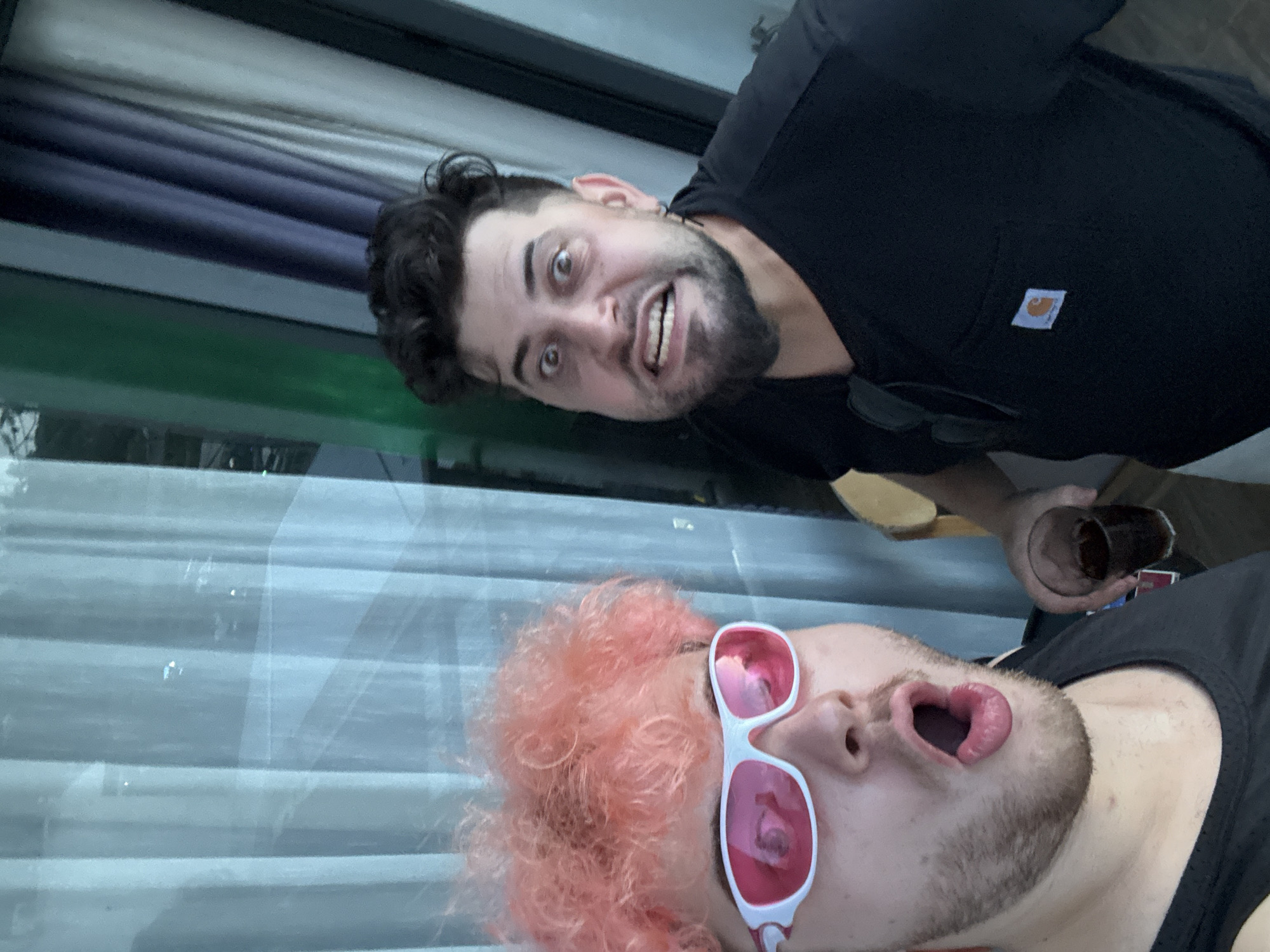 Two individuals are captured in a selfie on a balcony in Da Nang, Vietnam. The person in the foreground, on the left, has bright pink, curly hair and is wearing white sunglasses with pink lenses and a black tank top. Their mouth is open in an expression of surprise or excitement. The second person, on the right, has dark hair and a beard, wearing a black t-shirt with a Carhartt logo on the pocket. They are making a distorted facial expression with wide eyes and gritted teeth. A glass with a dark beverage is visible in the hand of the person on the right. Behind them, vertical curtains in various shades of blue and grey are drawn, with a hint of greenery and city lights visible through them, suggesting an urban setting.