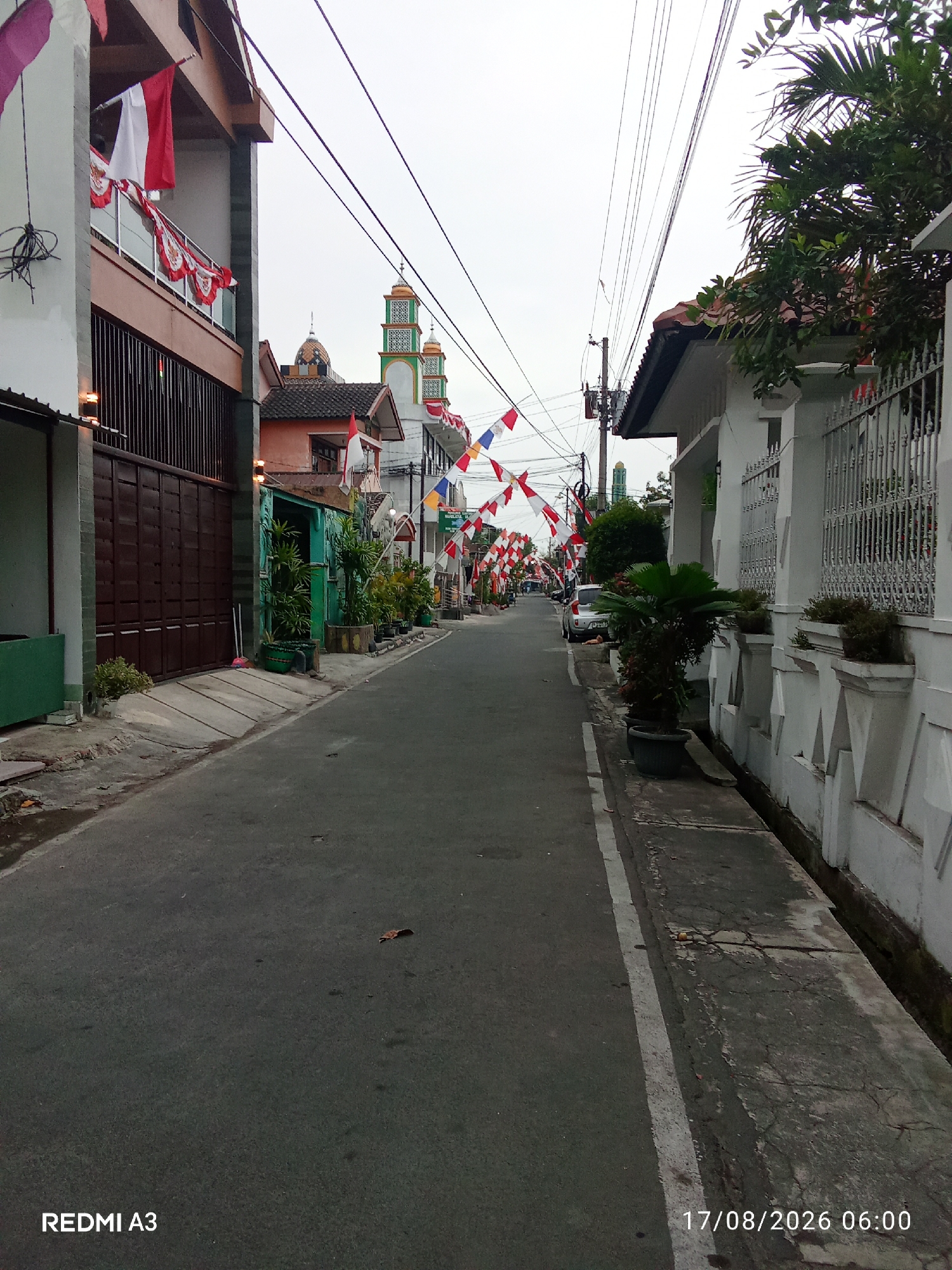 The image depicts a street scene in Banaran, Indonesia, adorned with red and white flags and pennants, suggesting a national celebration. The street is narrow and paved, with buildings lining both sides. On the left, there's a multi-story building with a large brown gate and balconies decorated with flags. Across the street, on the right, a white building with ornate fencing and potted plants is visible. In the distance, two mosque minarets with green and gold accents rise above the rooftops. A few parked cars and the presence of power lines indicate an urban or semi-urban environment. The sky is overcast, and the overall lighting suggests it is either early morning or late afternoon. The bottom of the image displays a timestamp "17/08/2026 06:00" and "REDMI A3".