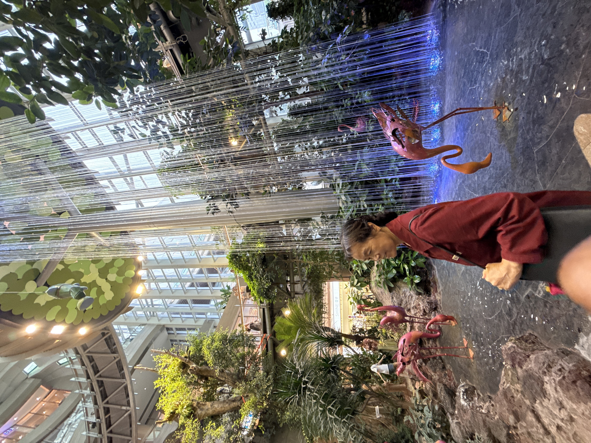 In the city of Liangjiang, China, a woman in a red jacket stands in the foreground, facing right, observing an indoor garden installation. The installation features a cascading water feature, illuminated with blue lights at its base, creating a curtain of falling water. Various tropical plants and trees are interspersed throughout the scene, creating a lush, green environment. Several decorative pink flamingos are placed around the water feature, some appearing to be submerged in the shallow water. In the background, multi-story buildings with large windows are visible, suggesting the installation is located within a large indoor complex, possibly a shopping mall or atrium. People can be seen on elevated walkways in the background, adding to the sense of scale and activity within the space. A circular, green-leafed structure hangs from the ceiling, adorned with lights and a small, green figurine suspended from it. The overall scene depicts a serene, nature-themed oasis within an urban architectural setting.