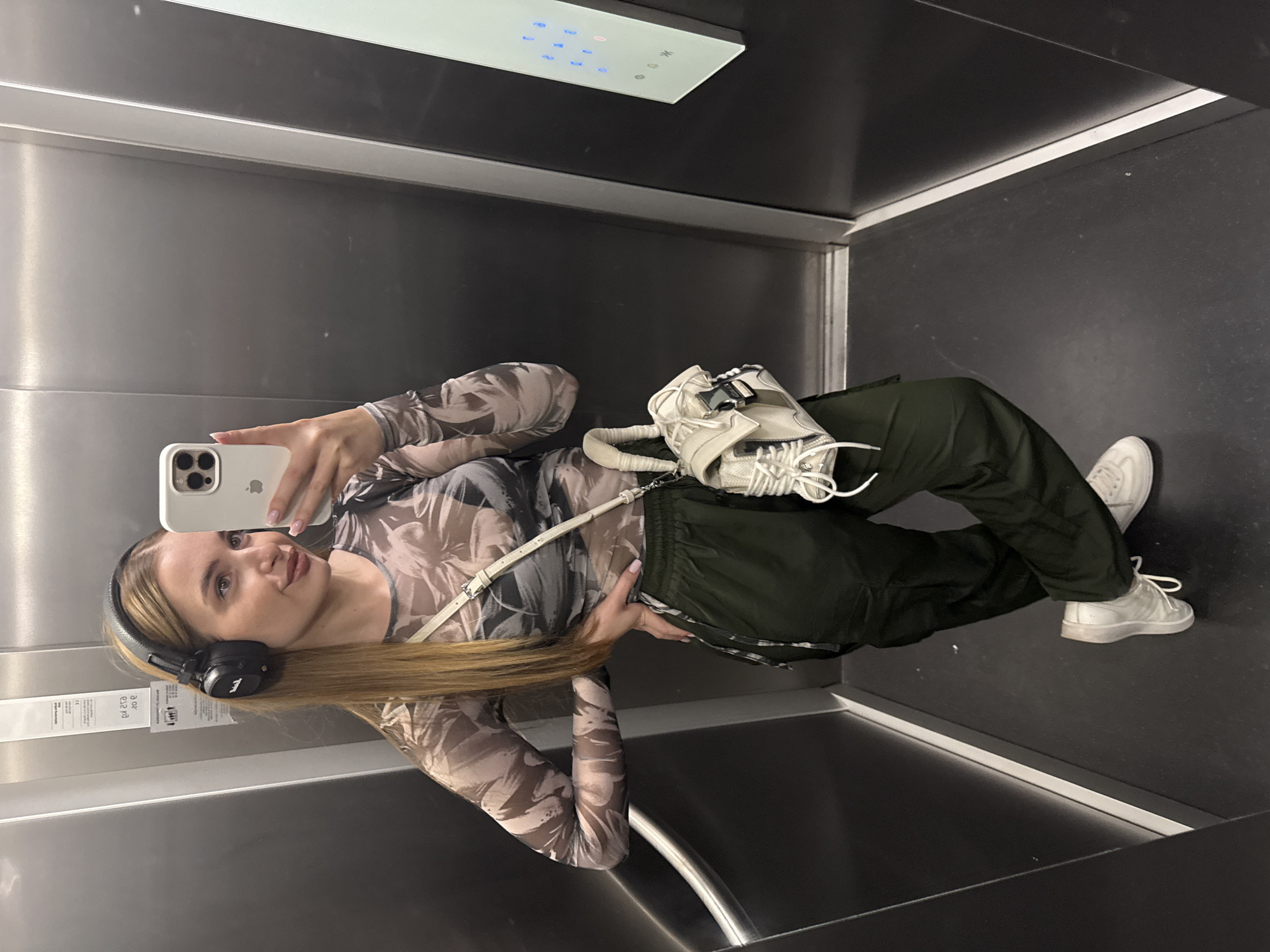 A blonde woman with headphones poses in an elevator, taking a photo in a mirror. She wears a patterned, long-sleeved top and dark green pants. She is wearing sneakers and a small white bag. The elevator has metal walls, a control panel at the top, and is likely located in an urban environment, but not specifically in Budva, Montenegro based on visual evidence.