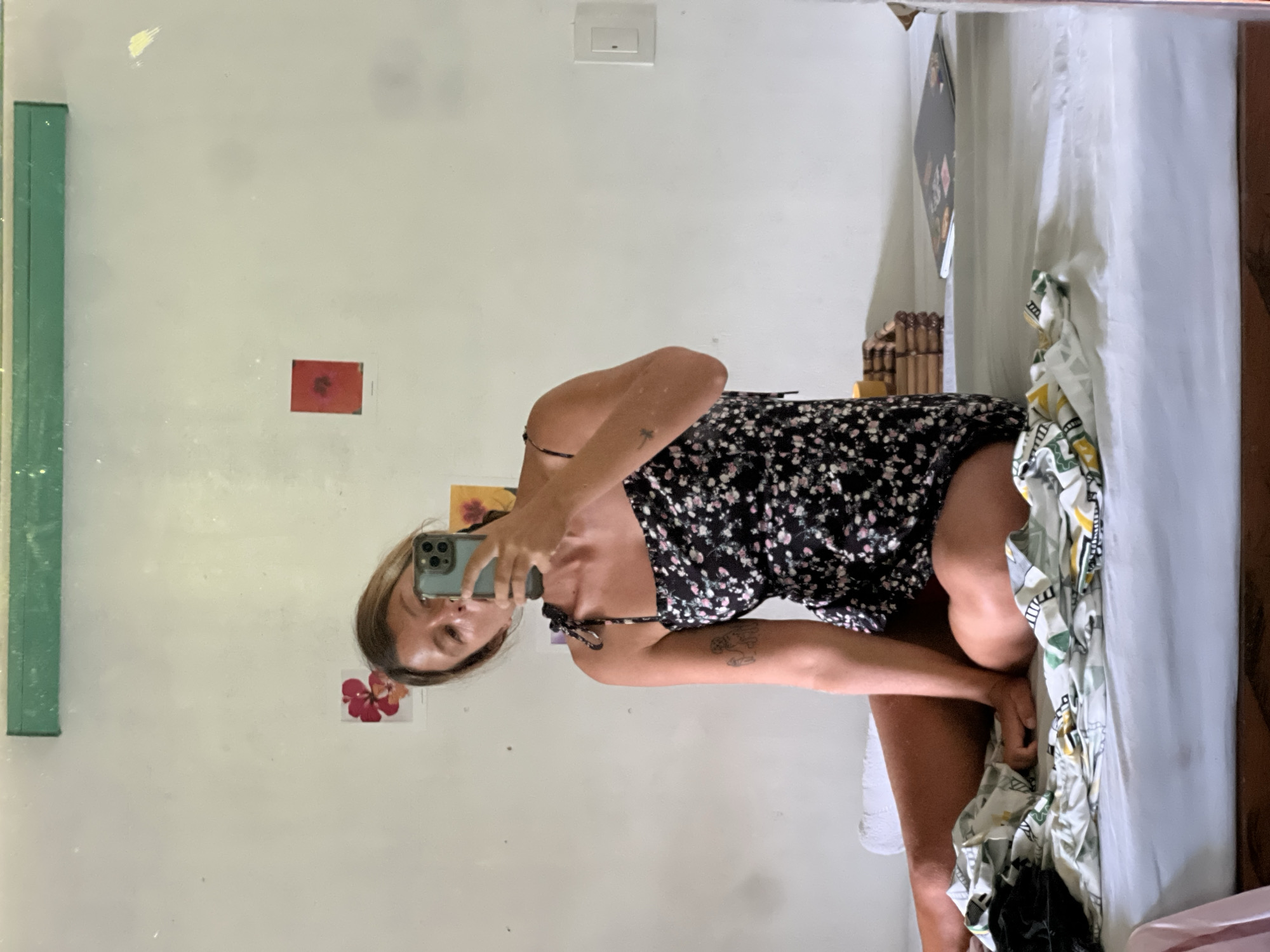 A woman is sitting on a bed and taking a mirror selfie. She is wearing a short black floral dress and has a phone in her right hand. The room has white walls, a green shelf above, and a window. There are some framed pictures and a white electrical outlet on the wall. The bed has a white sheet and a colorful, patterned duvet. The woman is in General Luna, Philippines.