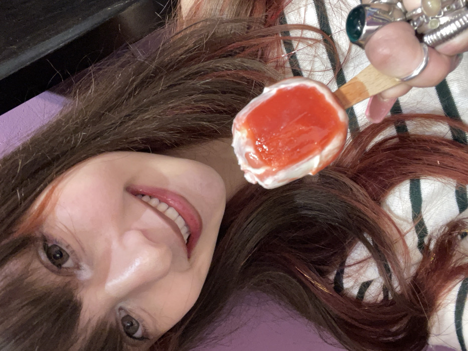 The image shows a close-up, high-angle view of a woman with long, dark brown hair, featuring noticeable reddish highlights, spread out around her head. Her face is angled upwards and to the left, revealing her left eye, nose, and a wide, open-mouthed smile that shows her white teeth. She has bangs covering part of her forehead.
Her right hand, adorned with multiple rings on several fingers (including a prominent silver-toned ring with a green stone on her index finger, and other rings with lighter stones), is visible in the upper right portion of the frame. This hand is holding a popsicle or ice cream bar on a wooden stick. The frozen treat has a white base, likely cream or vanilla flavored, generously topped with a bright, glossy red fruit puree, possibly strawberry or cherry, suggesting a summery or sweet indulgence.
The woman appears relaxed and happy, indicated by her joyful expression. Her hair is loosely spread, partially covering a striped garment (white with dark, possibly black or dark green, stripes) on the right side of the image, and resting against a solid purple surface on the left. A dark, possibly wooden or upholstered, surface is visible along the top edge of the frame. The overall impression is one of a candid, cheerful moment, perhaps enjoying a treat in a comfortable indoor setting. No text is visible in the image.