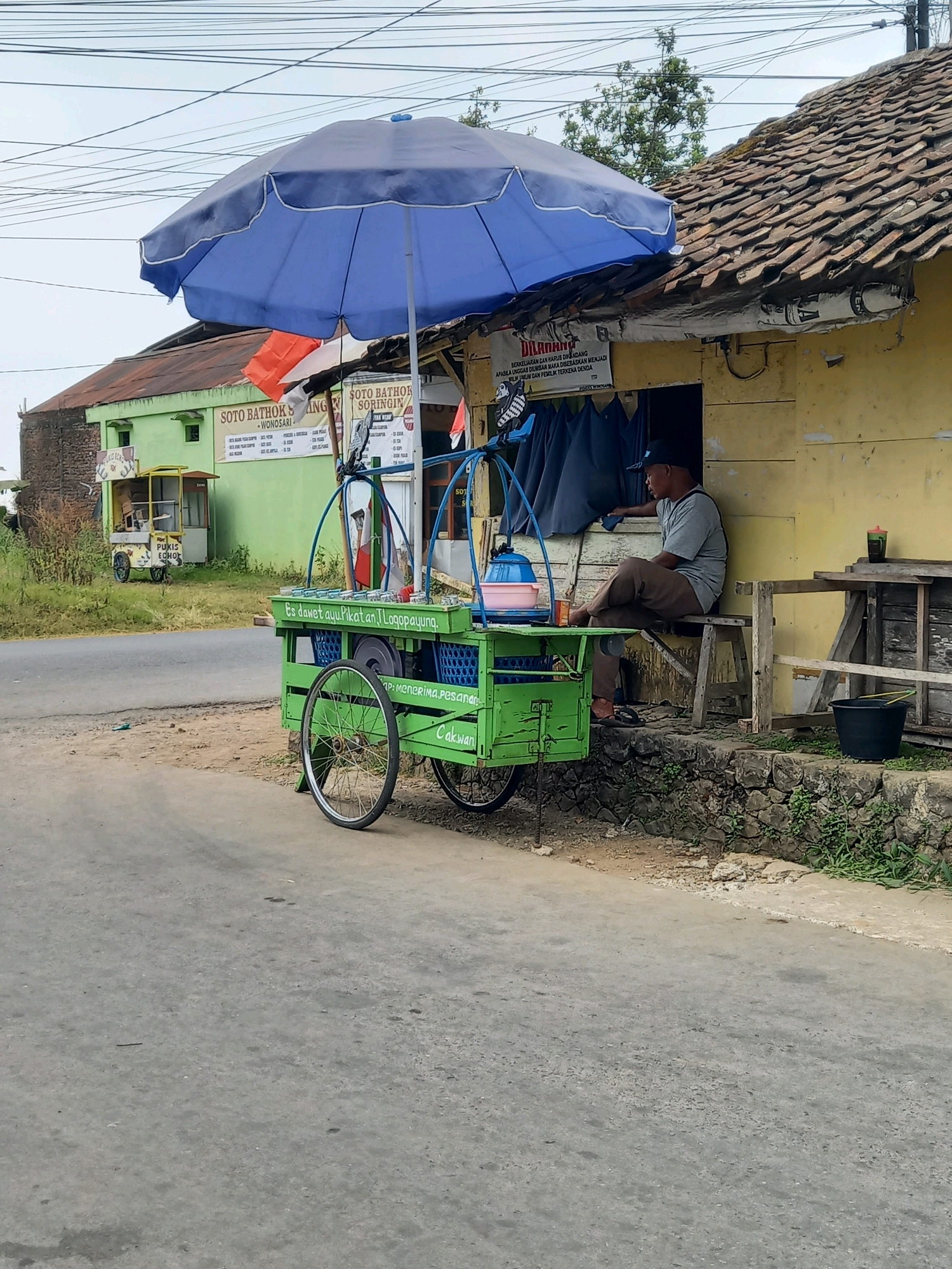 In Sangubanyu, Indonesia, a street vendor sits on a bench outside a yellow-painted shop with a corrugated tile roof. A blue umbrella is propped above a green cart filled with containers and items. The cart has "ES DAWET AYU PIKAT AN LOGOYOPAYUNG" written on it. A man wearing a grey t-shirt, brown pants, and a dark baseball cap sits on the bench, looking towards the shop entrance. Behind the shop, a green building with a sign that reads "SOTO BATHOK SRINGIN" is visible. The scene is set beside a paved road with a stone wall and vegetation on the right side.