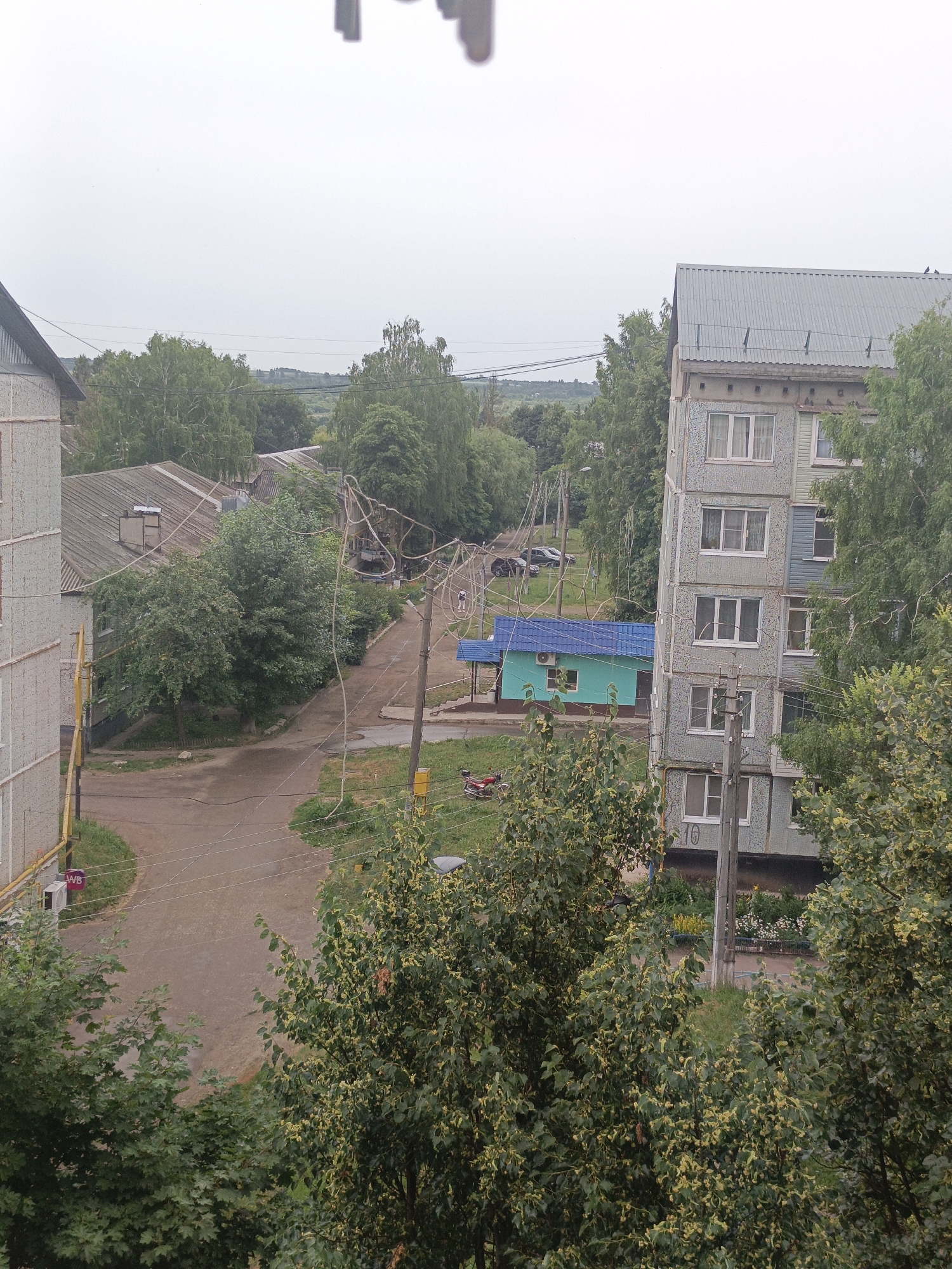The image shows a residential area with several buildings and trees. A multi-story apartment building is visible on the right, with multiple windows and light blue siding. To the left, another apartment building with a grey facade and white trim is partially visible. In the center of the image, a small turquoise building with a blue roof stands next to a dirt road. A few cars are parked on the side of the road. Trees with green foliage are scattered throughout the scene, with some flowering trees adding yellow accents. Utility poles and wires are present, indicating the presence of infrastructure. The sky is overcast. The scene appears to be a typical residential neighborhood in a city.