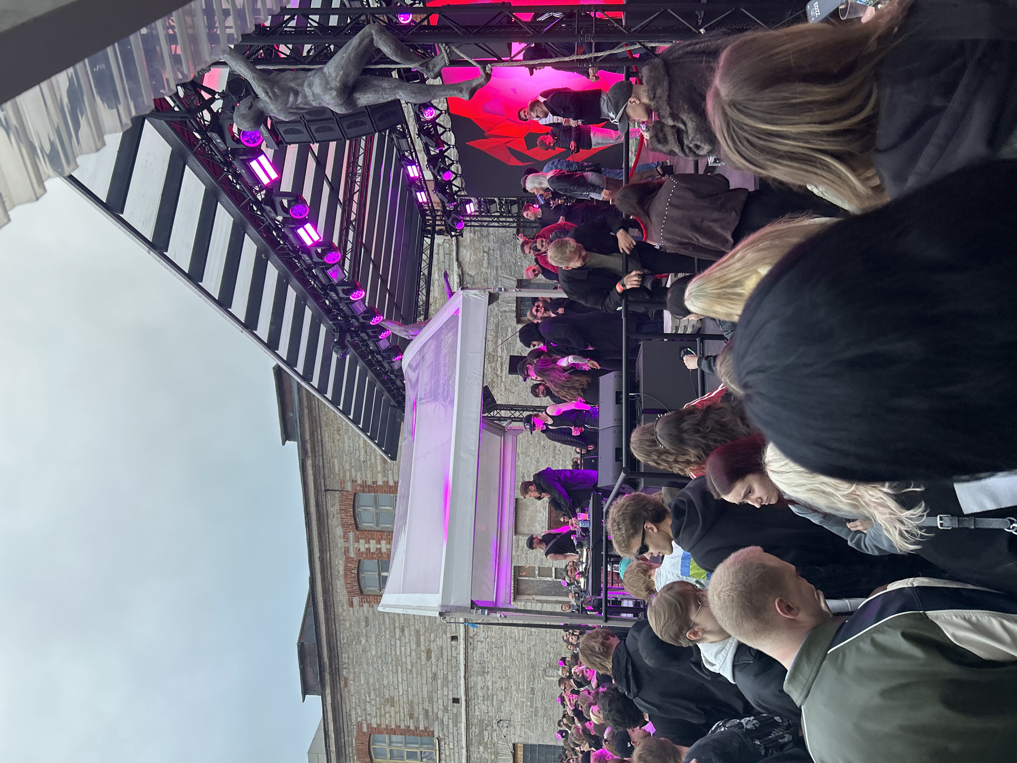 A large crowd of people are gathered outdoors in front of a stage setup at an event in Tallinn, Estonia. The stage has a large canopy, a screen displaying red graphics, and numerous pink stage lights. A statue of a person is mounted on a pole on the right side of the stage, its arm reaching up towards the canopy. Several individuals are on or near the stage, some appearing to be performers or staff, interacting with equipment that includes turntables and microphones. The audience is densely packed, with individuals of varying ages and styles of dress. In the background, a stone building with visible brickwork and windows is situated behind a smaller, white-canopied structure. The overall scene suggests an outdoor concert or festival.