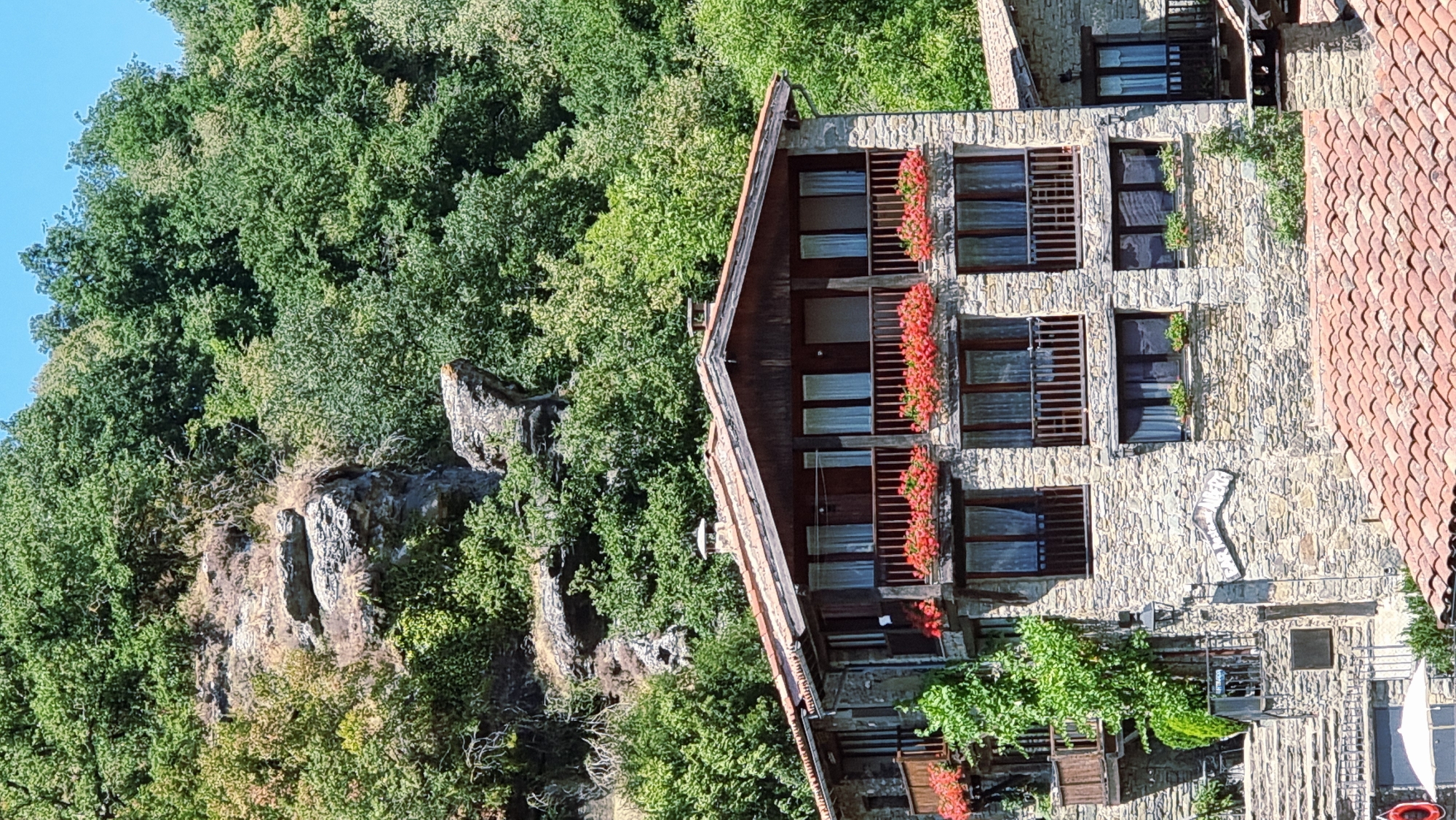 The image shows a stone building with a brown tiled roof, situated on a hillside in Rupit i Pruit, Spain. The building has multiple levels, each featuring balconies with wooden railings and red flowers in planters. Windows with white curtains are visible on each level. A lush green forest with a rocky outcrop serves as the backdrop. The sky is clear and blue. There are no people visible in the image.