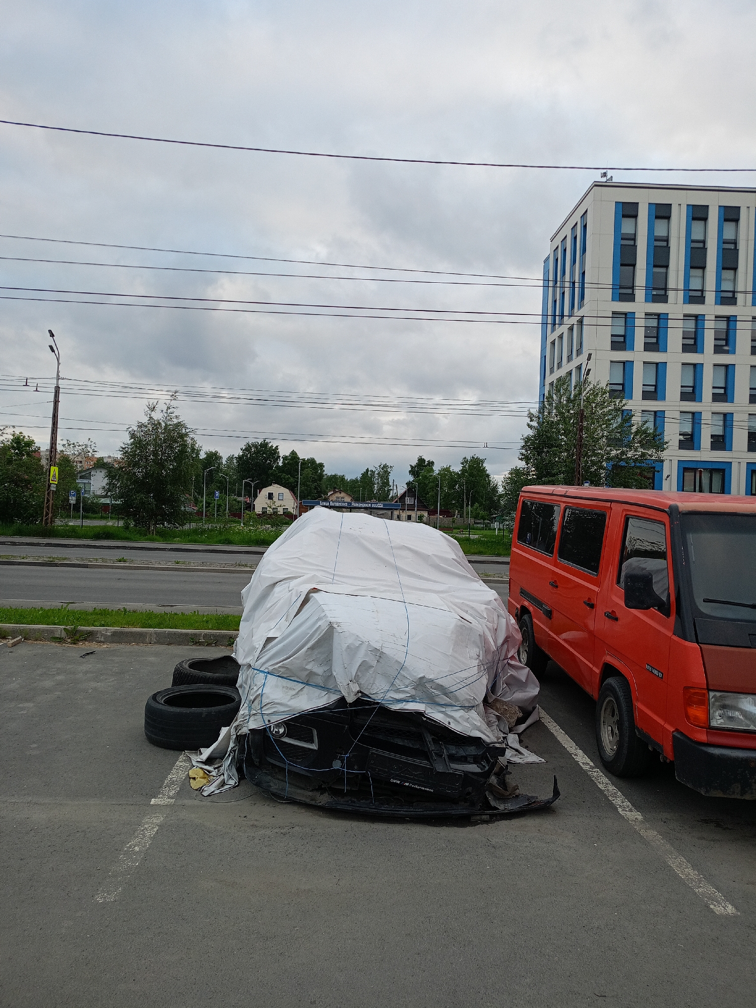In Petrozavodsk, Russia, on an overcast day, a damaged black car, partially covered by a white tarp secured with blue ropes, is parked in a lot. Adjacent to it is a bright orange van. The car appears to have significant front-end damage, with parts of the bumper and grill exposed. Two tires are placed beside the front of the car. In the background, a modern multi-story building with white and blue accents stands against a cloudy sky. A street with passing cars and a grassy median separates the parking lot from a row of trees and houses. Power lines are visible crisscrossing the sky.