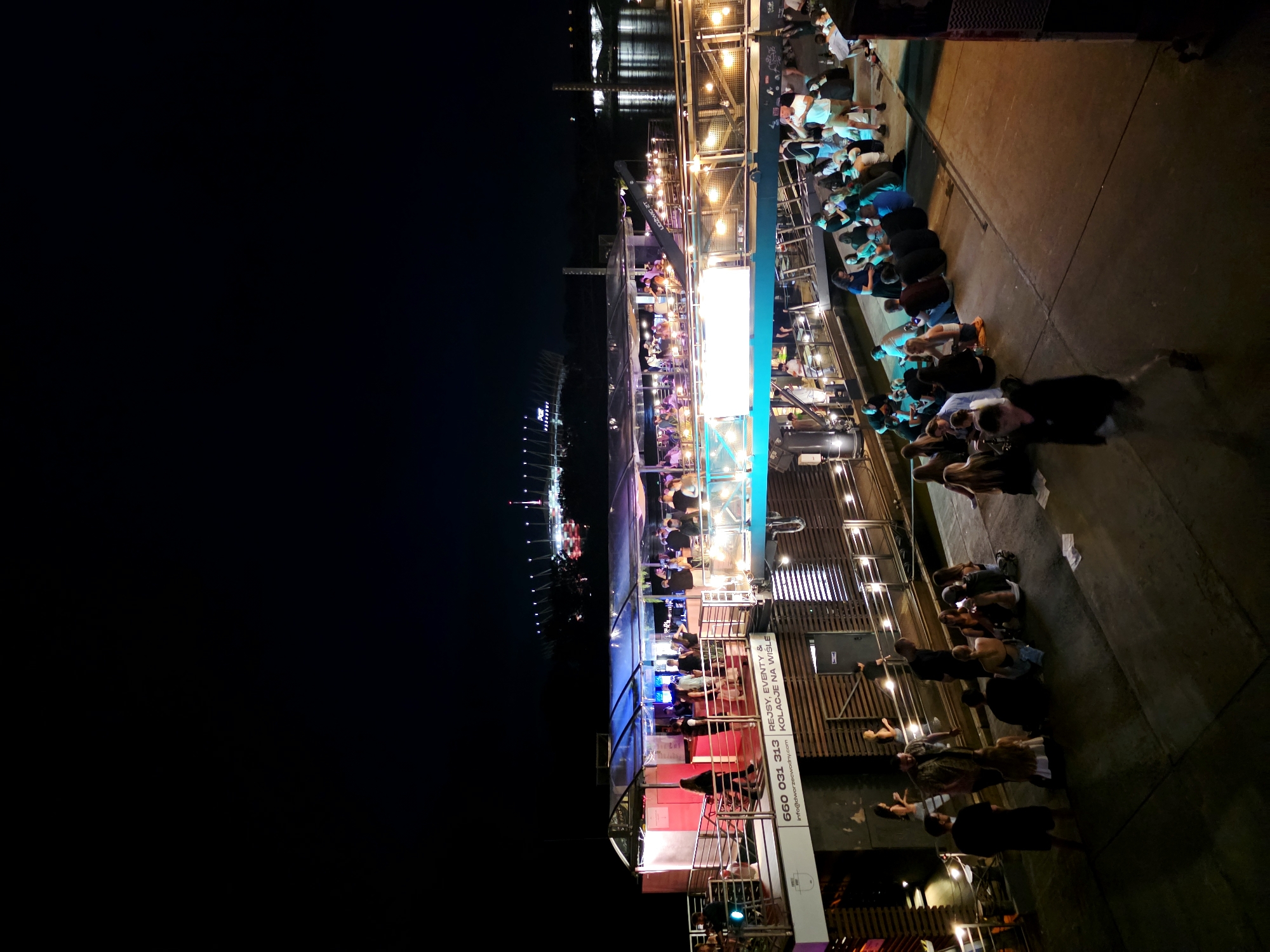 At night, a crowd of people gathers on a concrete embankment next to a multi-level venue on the Vistula River in Warsaw, Poland. The venue, which appears to be a bar or club on a boat or floating structure, is illuminated by various lights, including neon and string lights. People are seated and standing in groups on the embankment, some facing the river and others facing the venue. The upper levels of the venue are filled with patrons, and a bar area is visible. In the distance, across the river, a large illuminated stadium is visible, likely the PGE Narodowy stadium. The sky is dark, indicating it is nighttime.
