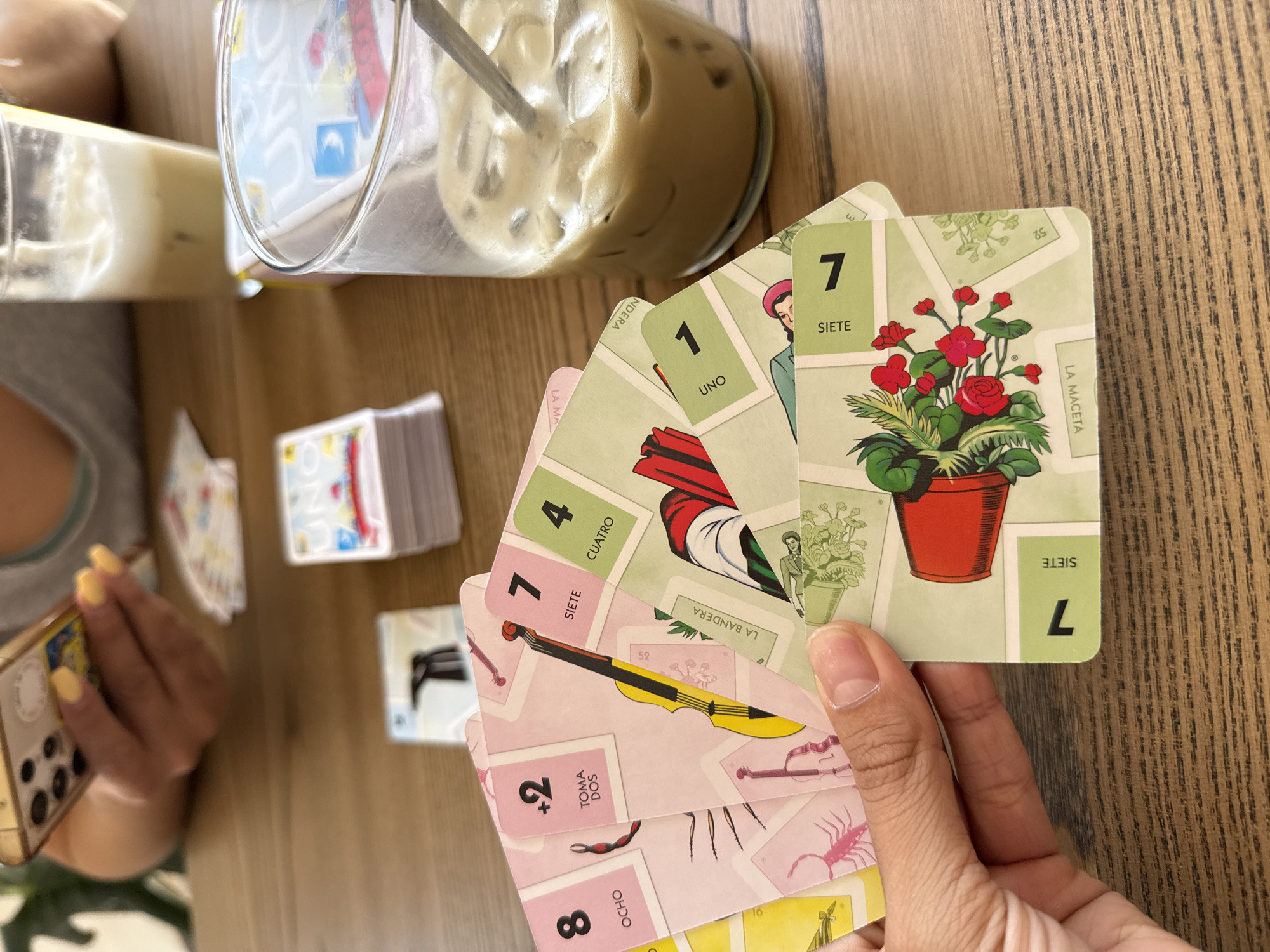 A person sits at a wooden table holding a hand of Lotería cards, a traditional Mexican game of chance, with coffee beverages in the background. The top card features a potted plant ("La Maceta"), followed by "La Bandera" (the flag), "Cuatro" (four), and others with Spanish terms and numbers. A separate stack of Lotería cards sits nearby. The person holding the cards is also holding a cell phone in their left hand. The scene is likely in a café or home setting. It is not possible to determine the location in the city of San Bernardino Tlaxcalancingo, Mexico, without additional information.