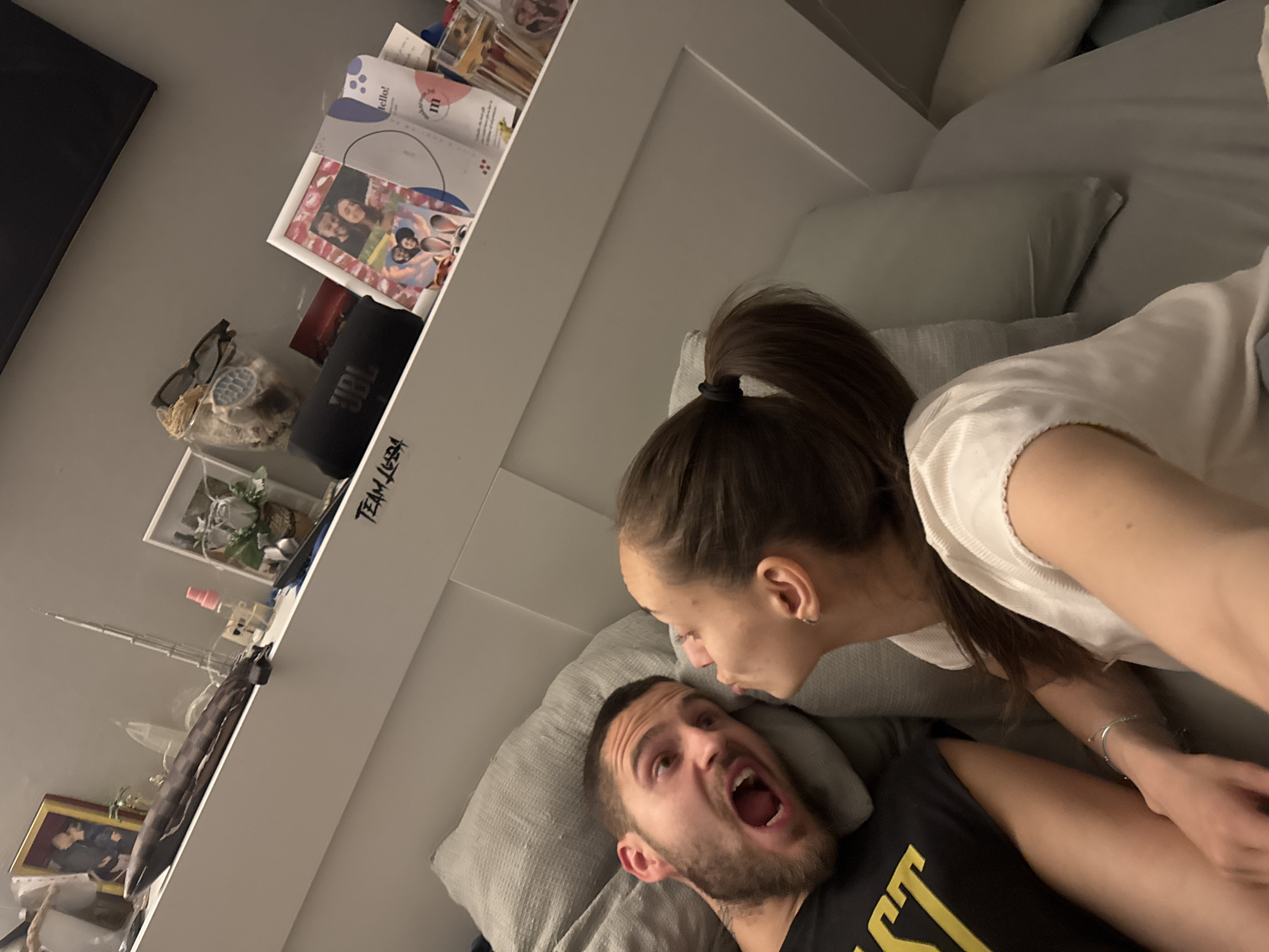 This indoor, tilted photograph captures a playful and intimate moment between a man and a woman, likely in a bedroom or living room in Sofia, Bulgaria. The woman, with long brown hair tied in a high ponytail and wearing a white top, leans over a man who lies on gray pillows. Her face is close to his, suggesting a teasing or affectionate interaction. The man, with a beard and wearing a dark tank top with yellow text, has his mouth wide open in a surprised or mock-shocked expression, conveying a lighthearted emotion.
The setting features a cozy home environment. Behind the individuals, a white headboard or shelf unit is visible, adorned with various personal items. These include multiple photo frames displaying pictures of people, a black JBL portable speaker, sunglasses, and decorative objects such as small plants, a glass jar containing shells or other trinkets, and a miniature tower-like sculpture. Books or magazines are also stacked on the shelf, one prominently displaying "hello!" and an "m" logo. A black screen, possibly a television, is partially visible on the far left.
The lighting indicates an indoor setting, potentially evening or night, given the absence of strong natural light. Visible text includes "JBL" on the speaker, "hello!" and an "m" logo on a magazine, and the word "BEST" partially visible in yellow letters on the man's tank top. Additionally, the phrase "TEAM JGTH" (or similar script) is written vertically in black marker on the side of the white shelf. The overall mood of the image is casual, playful, and affectionate.