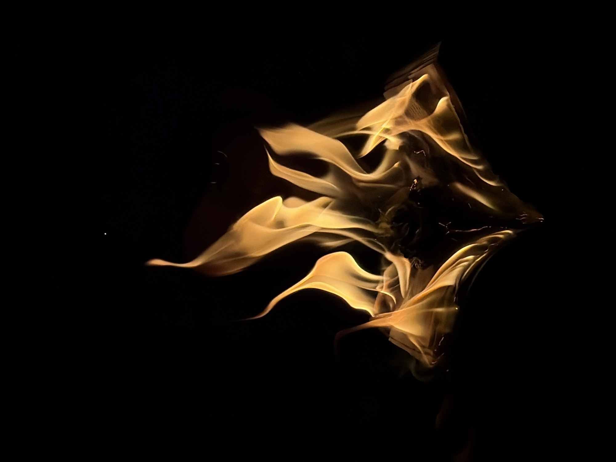 The image depicts yellow flames in the darkness. The shape of the flame is abstract and complex. The background is entirely black. There is no indication of it being in Chaikovsky, Russia, or any specific location. There are no people, objects, or actions visible other than the flames themselves.