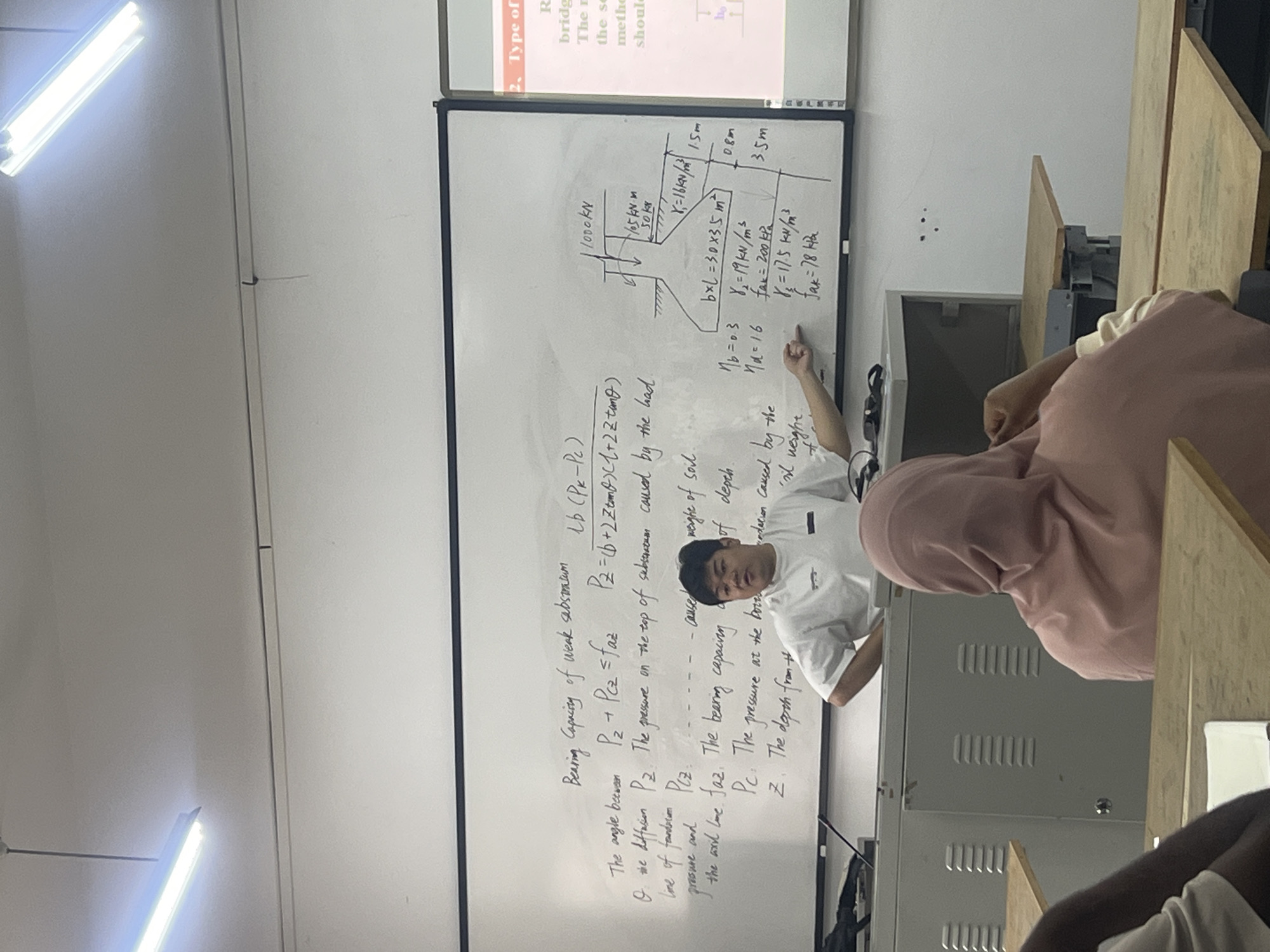 The image shows a classroom setting, potentially in Jiangjin, China, though this is based on context clues rather than direct evidence in the image. A whiteboard displays a diagram of a bridge structure with labeled measurements and bearing capacity equations. A person is pointing to the board, likely a student presenting or an instructor teaching. Another student is seated, listening to the presenter. The whiteboard also shows bearing capacity calculations.