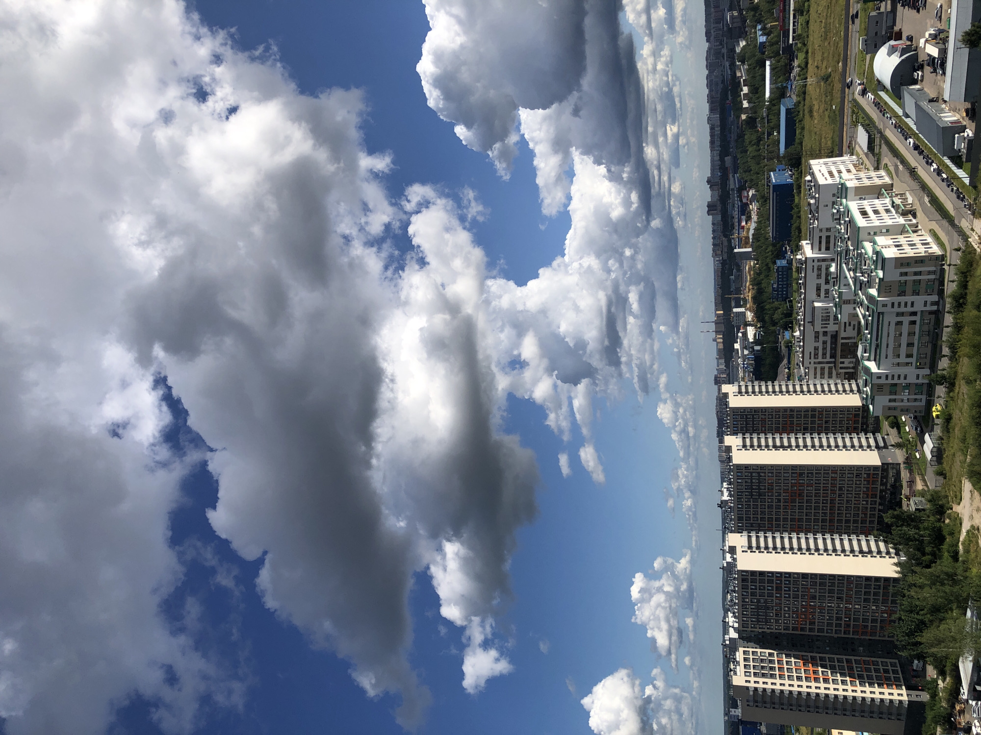 An aerial view captures a vast urban landscape under a dramatic sky filled with an array of cumulonimbus and stratocumulus clouds. The foreground is dominated by several large, modern apartment buildings. The most prominent are a cluster of interconnected high-rise buildings with a striking facade of dark grey, cream, and orange panels, featuring numerous windows. To the right and further back, other apartment complexes with white and green accents are visible. The buildings are surrounded by patches of greenery, including trees and grassy areas, and a network of roads with parked cars. In the distance, the urban sprawl continues with more buildings, industrial structures, and a line of trees marking the horizon. The sky is a vibrant blue, with sunlight illuminating the white portions of the clouds and casting shadows on the darker, denser parts, creating a dynamic interplay of light and shade. The overall impression is of a contemporary city on a day with unsettled weather.