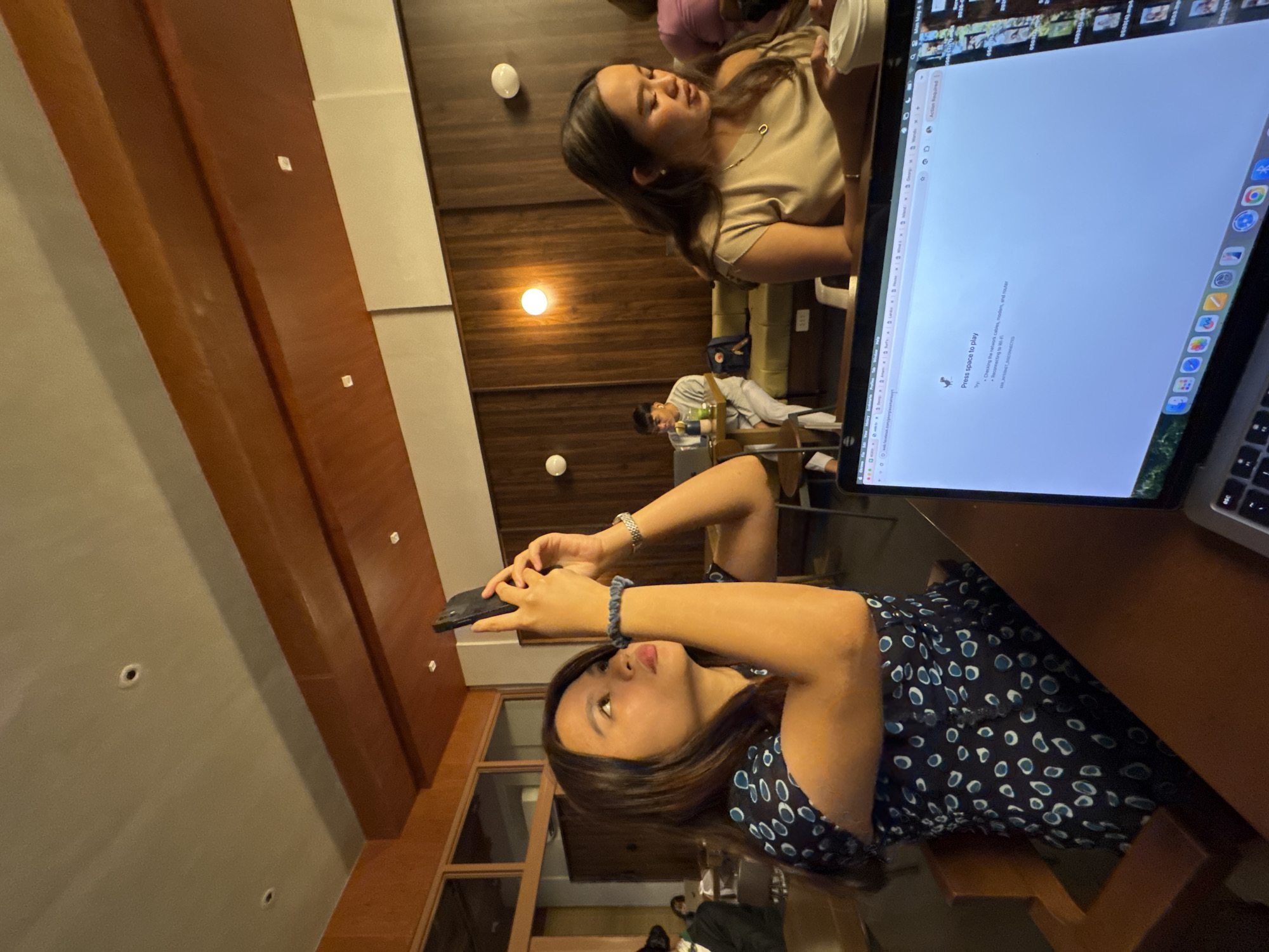 The image shows an indoor scene at what appears to be a café or co-working space. A woman in a dark blue dress with a circular pattern is holding a smartphone and looking upwards, possibly taking a picture or video. Next to her is an open laptop displaying a "Press space to play" message, suggesting there is a network connectivity issue. Another woman is seated nearby, holding a beverage container and looking towards the laptop screen. In the background, a man is seated at another table, also using a laptop. The environment has wooden walls and ceiling details, and there are other people and objects visible in the blurred background, indicative of a public place. Given the location information provided in the prompt, this scene is likely taking place somewhere in Davao City, Philippines.