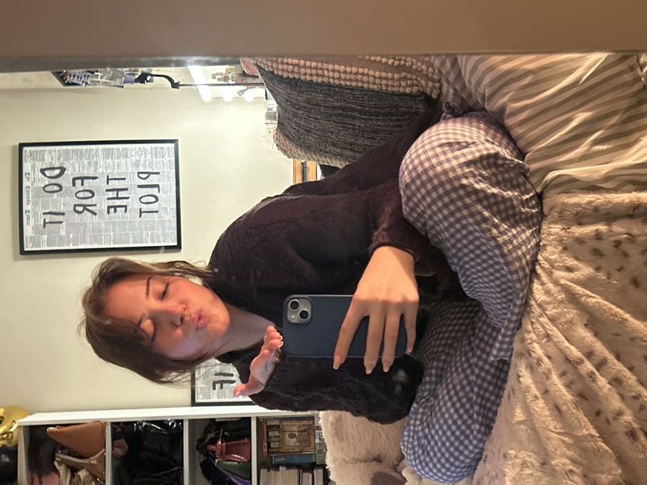 A young woman in a dark sweater and blue and white checkered pajamas takes a selfie. She puckers her lips and holds her phone in her right hand. She sits on a bed with multiple pillows and blankets. Behind her is a white storage unit with items inside, and a framed poster with text "DO IT FOR THE PLOT" on the wall. The image appears to be a mirror reflection.
