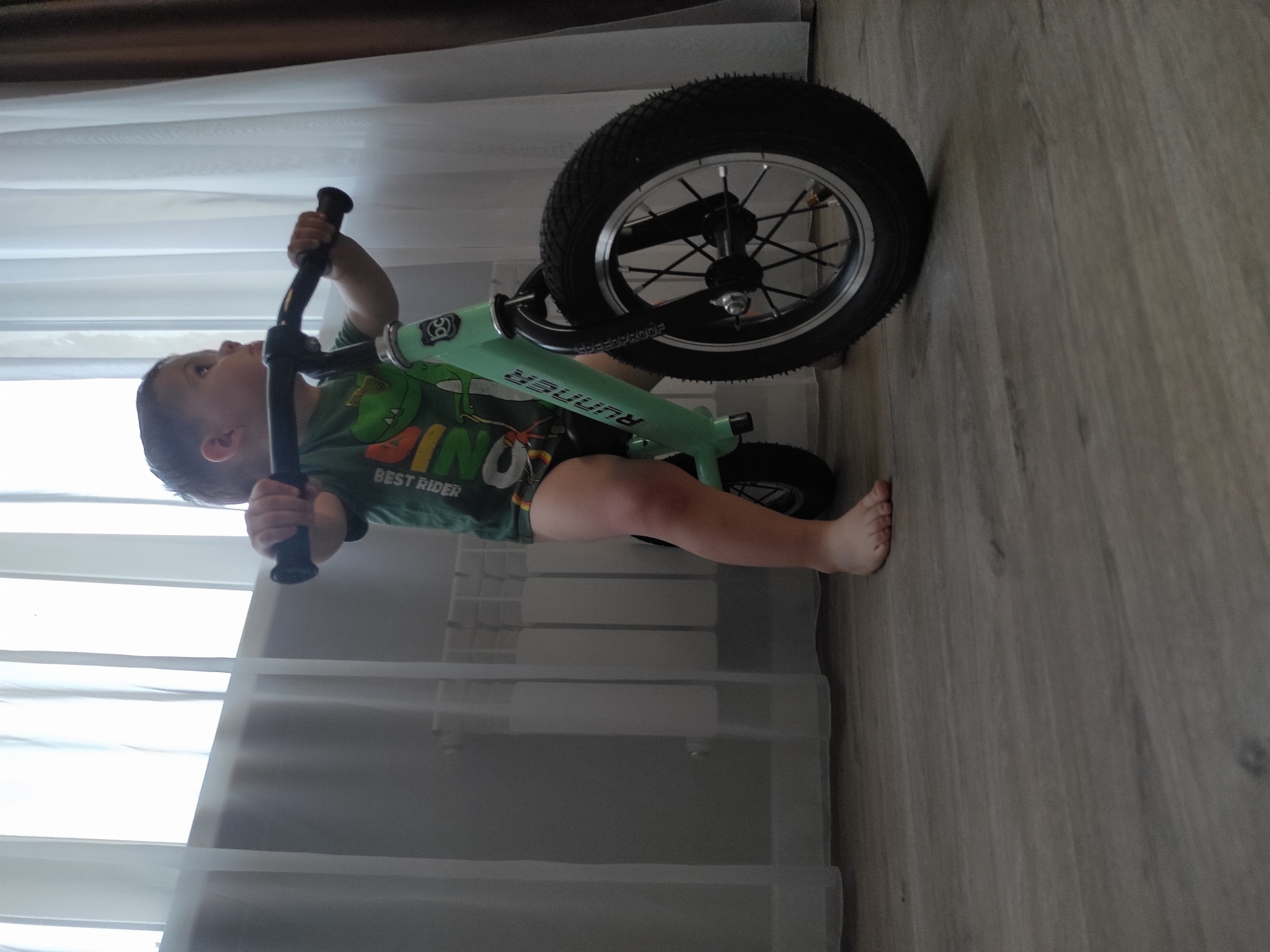 The image shows a young child, likely a toddler, on a green balance bike. The child is wearing a green t-shirt with a dinosaur graphic and the words "DINO" and "BEST RIDER" visible. The child is holding onto the handlebars of the bike. The bike has a large black tire with silver spokes and a black rim. The scene is indoors, with light-colored flooring that appears to be wood or laminate. In the background, there are white curtains and a radiator. The lighting suggests it is daytime, possibly late morning or afternoon, and the environment appears to be a home. The child's expression is not clearly visible, but the overall impression is of a playful and active moment. No specific weather or time of day is discernible beyond it being daylight.