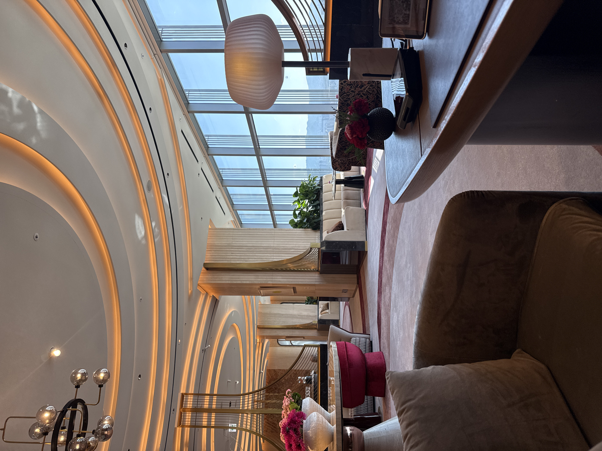The image captures an interior shot of a luxurious lounge area, likely within a hotel or upscale building. The location is identified as Seoul, South Korea. The lounge features a unique architectural design with curved walls and illuminated recesses, complemented by a large glass window providing ample natural light and a view of the cityscape. The decor includes a mix of modern and classic elements, such as a contemporary light fixture, plush seating arrangements, and decorative flower arrangements. The overall atmosphere suggests a high-end establishment with a focus on aesthetics and comfort.