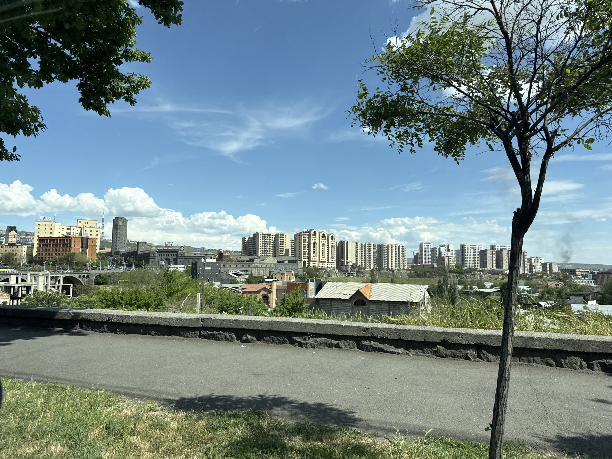 The image captures a daytime view of Yerevan, Armenia, showcasing a cityscape under a partly cloudy sky. In the foreground, a paved road runs parallel to a low stone wall. Grass grows along the edge of the road and below the wall. To the left, a tree branch with green leaves frames the scene. On the right, a slender tree trunk with sparse foliage stands prominently.
In the middle ground, buildings of varying heights and architectural styles populate the cityscape. A mix of residential and commercial structures are visible. Some buildings have modern facades, while others appear older. In the background, light-colored, high-rise apartment buildings are arranged in a line, stretching along the horizon.
Smoke rises in the distance, potentially indicating industrial activity or localized burning. The sky is blue with scattered white clouds.