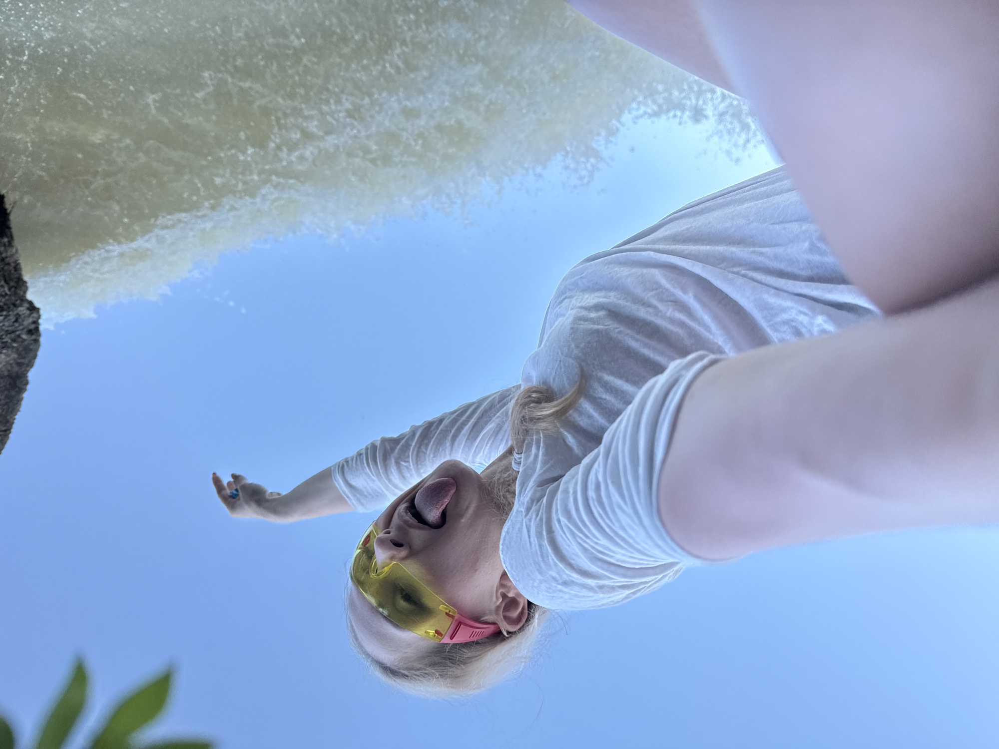 The image shows a woman lying on her back at the edge of a body of water, likely in Novyi, Russia. The water appears to be flowing over a small waterfall or dam. The woman is wearing a white shirt, yellow sunglasses, and a blue ring. She is sticking her tongue out and has her arm extended. The sky is visible in the background, with some greenery in the lower left corner.