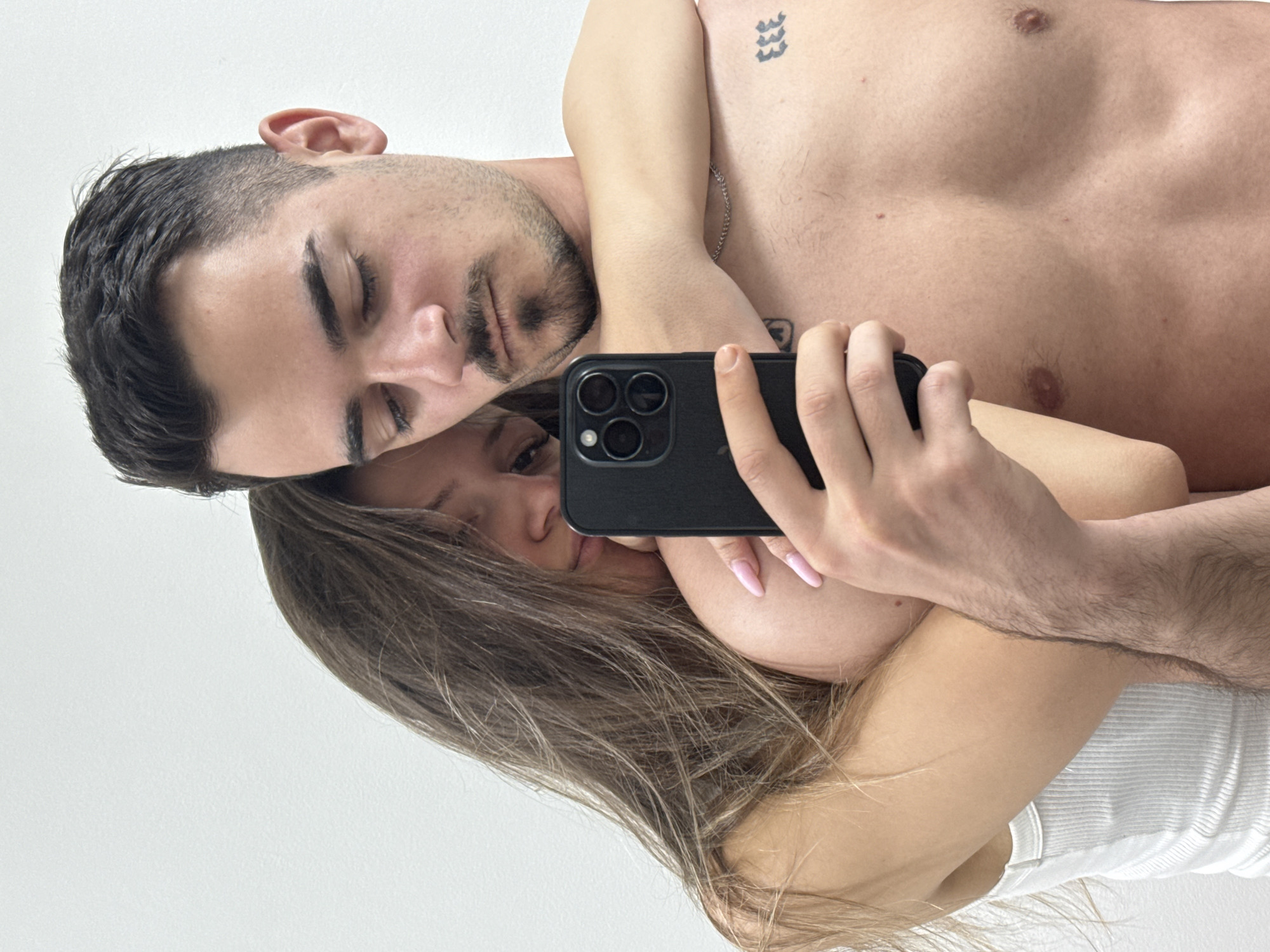 A photograph depicts a man and a woman in close proximity, taking a selfie. The man is shirtless with dark hair and closed eyes, displaying a tattoo on his upper chest. The woman, wearing a white tank top, holds a black iPhone with three camera lenses, capturing the image. The background appears to be a plain white wall. The location, Sofia, Bulgaria, cannot be confirmed from the visual information.