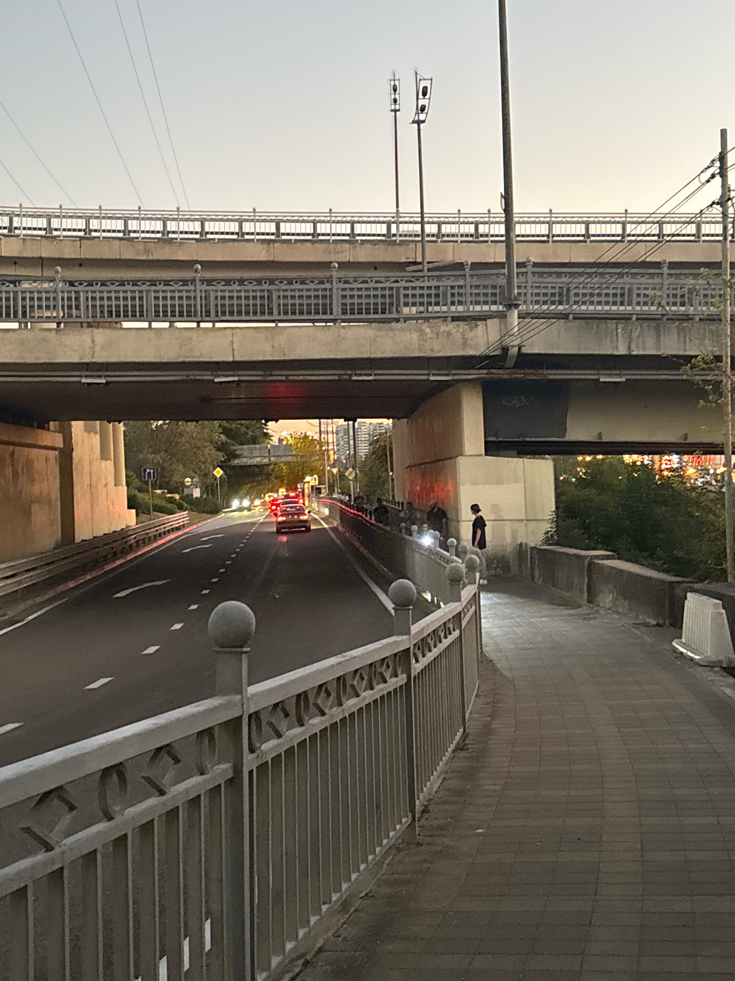 This image depicts an urban scene in Sochi, Russia, likely during twilight. A multi-tiered overpass dominates the upper portion of the frame, with concrete structures and railings visible. Below the overpass, a two-lane road is shown with cars driving, their headlights and taillights creating streaks of light. The road surface is asphalt with white lane markings. To the right of the road, a paved sidewalk runs alongside a decorative metal fence. Several individuals are present on the sidewalk, appearing to be standing and observing the scene. One person in a dark shirt stands out, facing away from the camera. The background reveals trees and buildings in the distance, with city lights beginning to illuminate the dusk. The overall atmosphere suggests a typical urban environment with infrastructure and pedestrian activity.