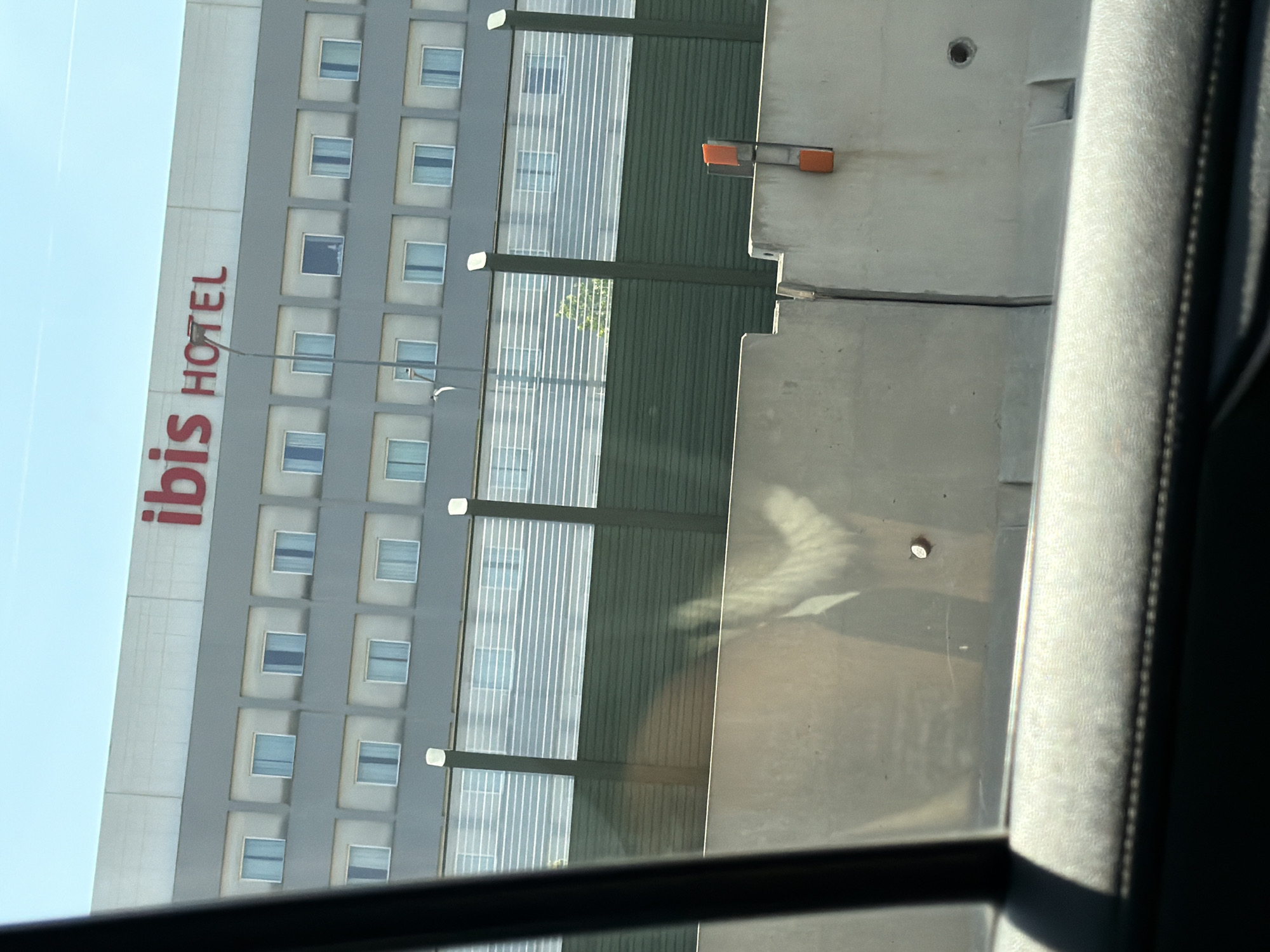 A shot taken from inside a vehicle looking out at the Ibis Hotel in Lainate, Italy. The hotel is a multi-story building with a grey facade and rows of windows. The words "ibis HOTEL" are visible in red lettering at the top of the building. In the foreground, there are concrete barriers and a reflective fence. The sky is clear and blue. The image appears to be taken during daylight hours.