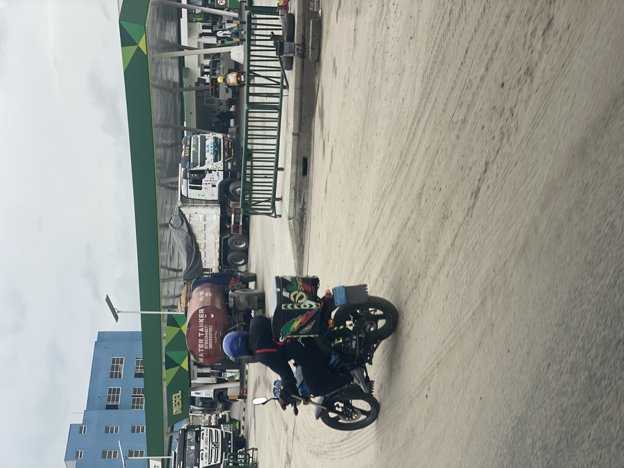 A medium shot of a gas station in Lekki, Nigeria. A motorcyclist wearing a blue helmet and dark clothing is riding away from the station on a paved road. The motorcycle has a storage box attached. A "WATER TANKER" is visible among the trucks, which include a white articulated semi-truck, under the gas station awning. The sky is overcast.