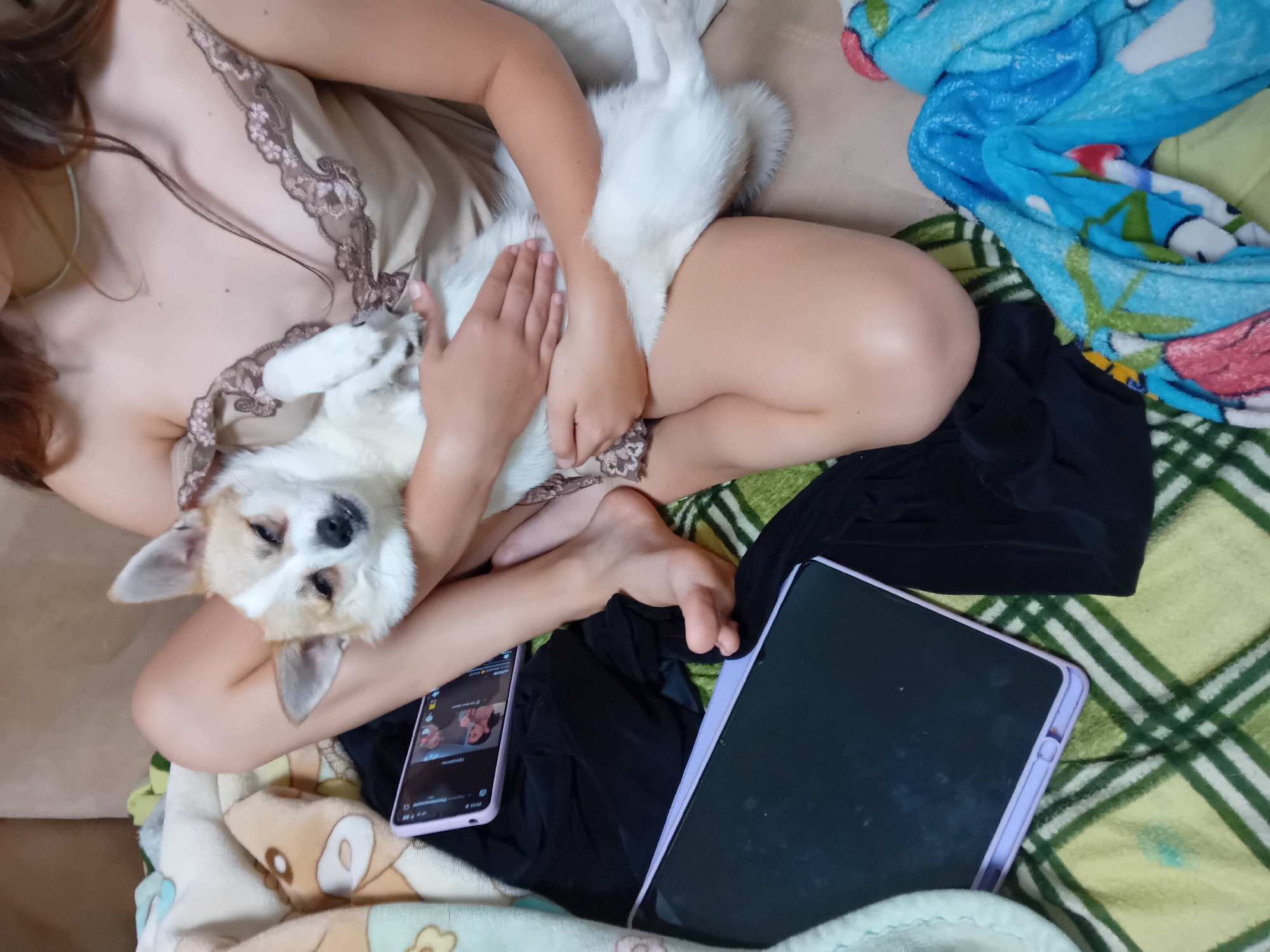 A person is holding a small dog in their lap while lying down on a sofa. The person is wearing a light-colored, lace-trimmed slip. The dog is white with tan markings around its face and ears and is looking towards the camera. There are several objects on the sofa, including a tablet, a smartphone, and blankets. One blanket has a green and yellow plaid pattern, while another is blue with cartoon characters. The setting appears to be indoors, likely a living room. There is no indication of the location in the city of Chuhuiv, Ukraine.
