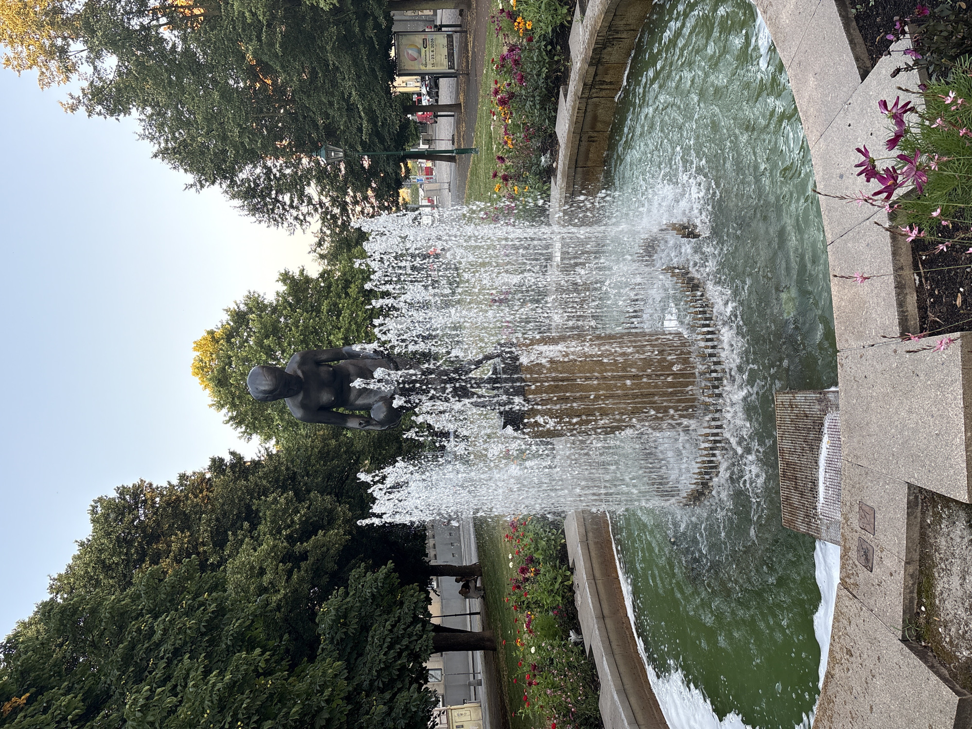 A bronze sculpture of a figure holding a sphere stands in the center of a circular fountain in Pilsen, Czechia. The fountain is surrounded by a low stone wall and a flowerbed filled with various colored flowers, predominantly pink and red. Water shoots upwards from the fountain, creating a curtain of spray around the sculpture. Behind the fountain, mature trees with lush green foliage line a walkway. In the background, a street is visible with parked cars and buildings. The sky is clear and pale blue, suggesting it is late afternoon or early morning. There are two people sitting on a bench under the trees in the mid-ground, and a lamppost is visible to the right.