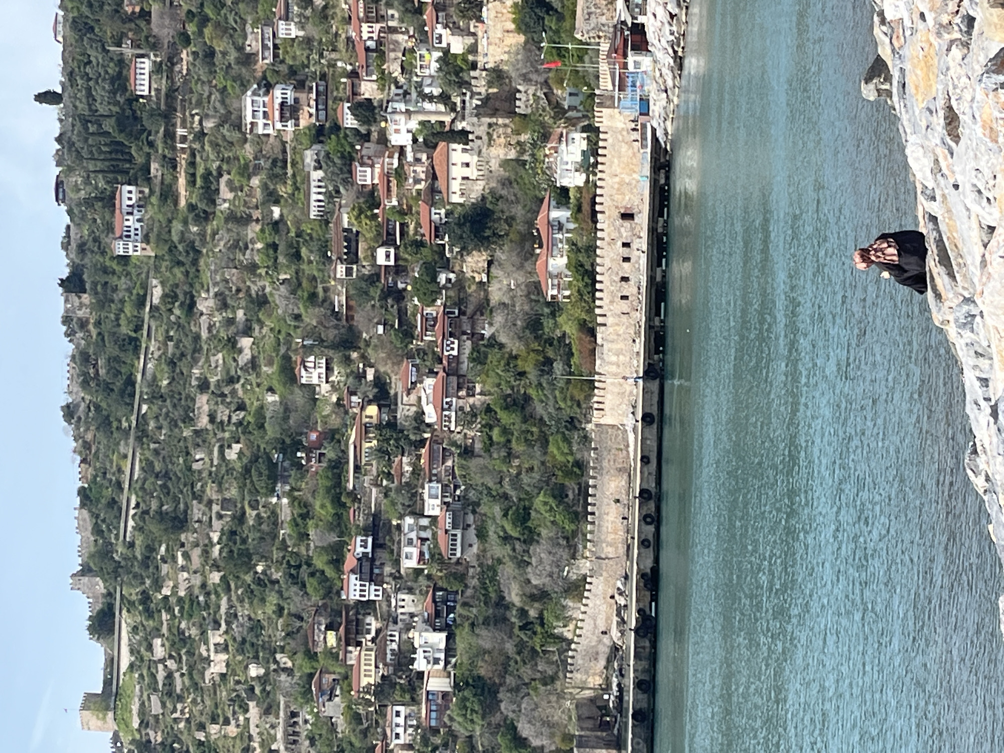 The image shows a scenic view from a high vantage point in Alanya, Turkey. It features a hillside densely populated with buildings and greenery, cascading down towards a body of water, likely the Mediterranean Sea. Along the waterfront, a stone fortification wall extends into the water, marked by regular crenellations and dark bollards lining its edge. The water appears calm and reflects the daylight. On the right side of the image, a person is visible, partially obscured, looking towards the view. The day appears bright with a clear sky.