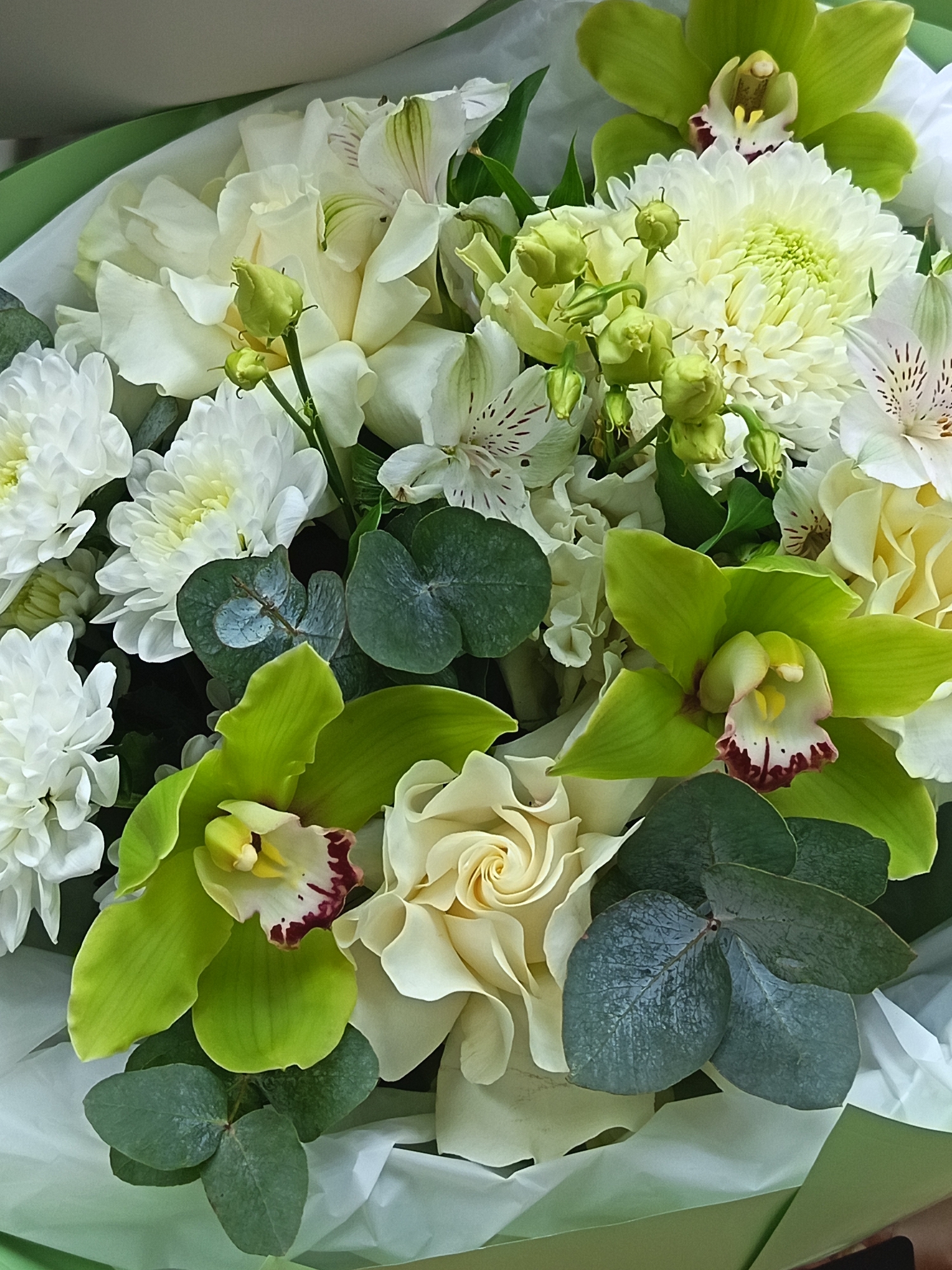 The image shows a close-up of a floral arrangement. Predominantly white chrysanthemums and alstroemeria are arranged alongside green orchids. Eucalyptus foliage is interspersed throughout the bouquet. A single, cream-colored rose is also present. The flowers are arranged in a package, presumably for gifting. There is no evidence of a specific location or the city of Nizhnevartovsk, Russia.