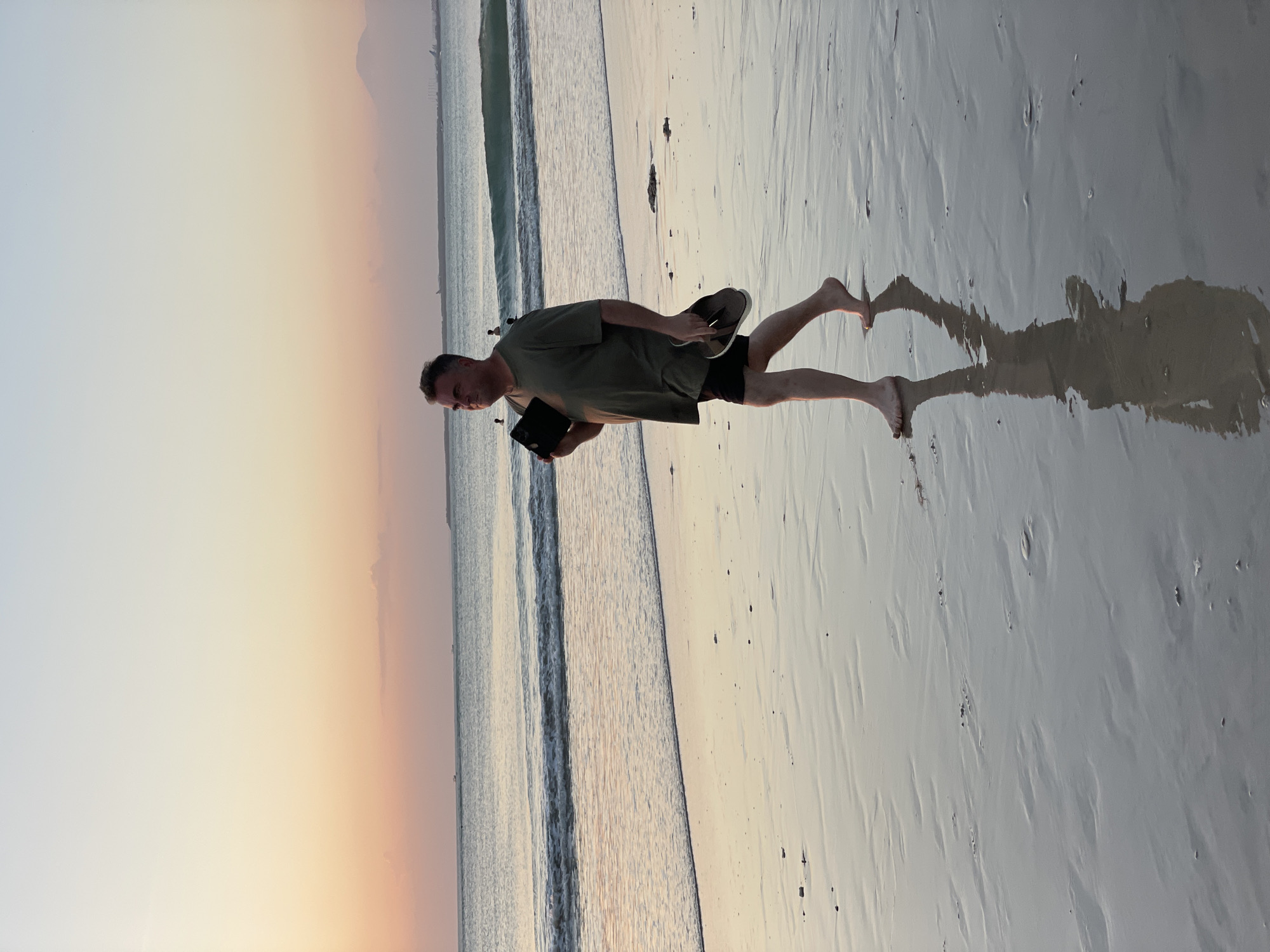 A man walks barefoot on a wet sandy beach during sunset. He is wearing a green t-shirt and black shorts and is holding a black tablet and a pair of flip-flops. The ocean is to his left, with gentle waves lapping at the shore. The sky is a gradient of pale yellow, orange, and pink, indicating the sun is setting. The wet sand reflects the sky and the man's shadow. In the background, there are faint silhouettes of ships and a distant landmass.