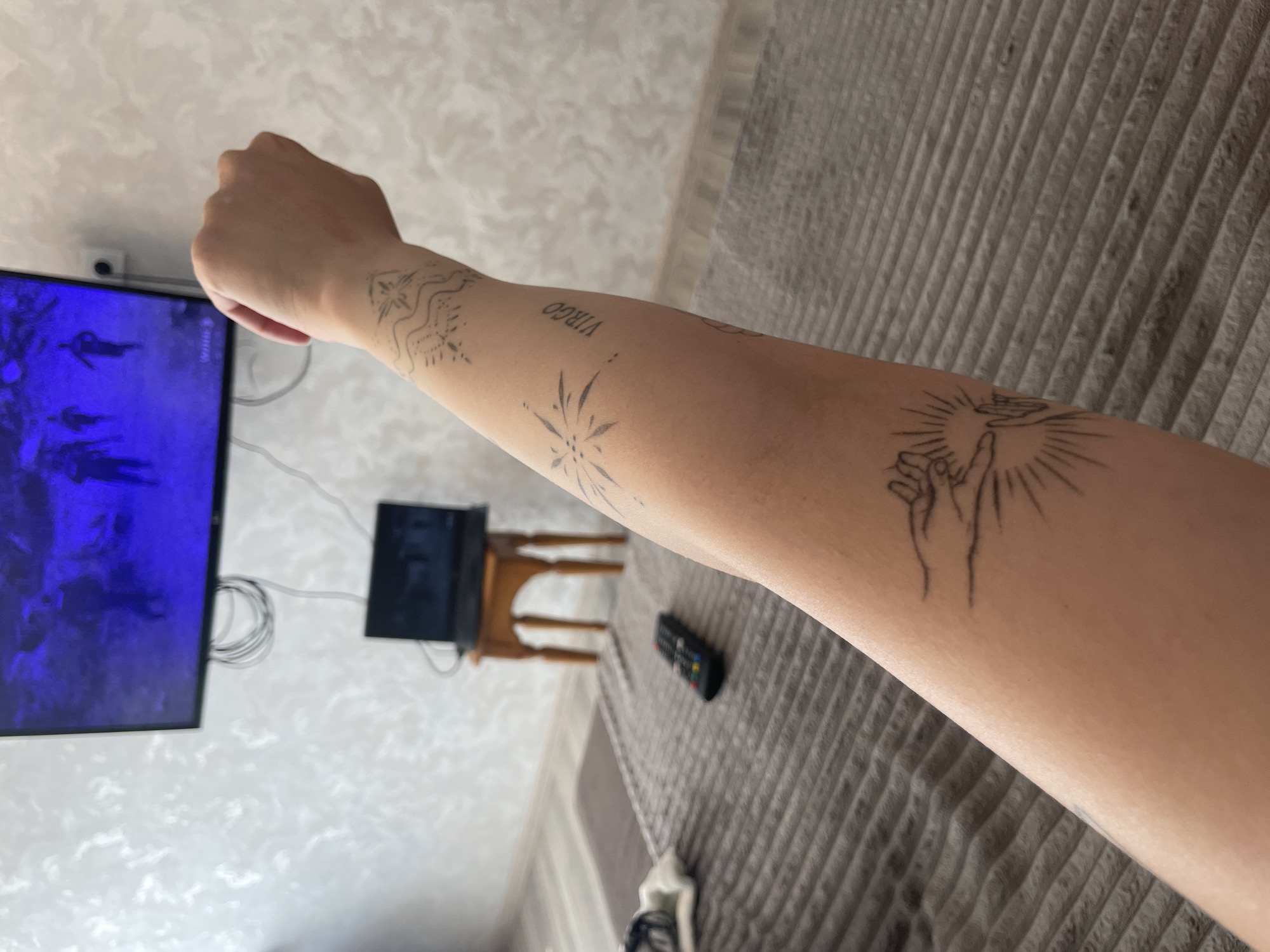 The image shows a close-up view of a person's arm with several line-drawn tattoos. The arm is positioned in front of a television screen displaying a dimly lit, possibly night-vision, image of figures in an outdoor setting. Below the television, a laptop is open on a small wooden table. The room appears to be a bedroom, with a bed covered in a gray, ribbed comforter in the foreground. A black television remote control rests on the comforter. The wallpaper in the background has a textured, possibly damask, pattern. The lighting is subdued, suggesting indoor illumination. The image does not contain information that would allow for identifying the location as Stavropol, Russia.