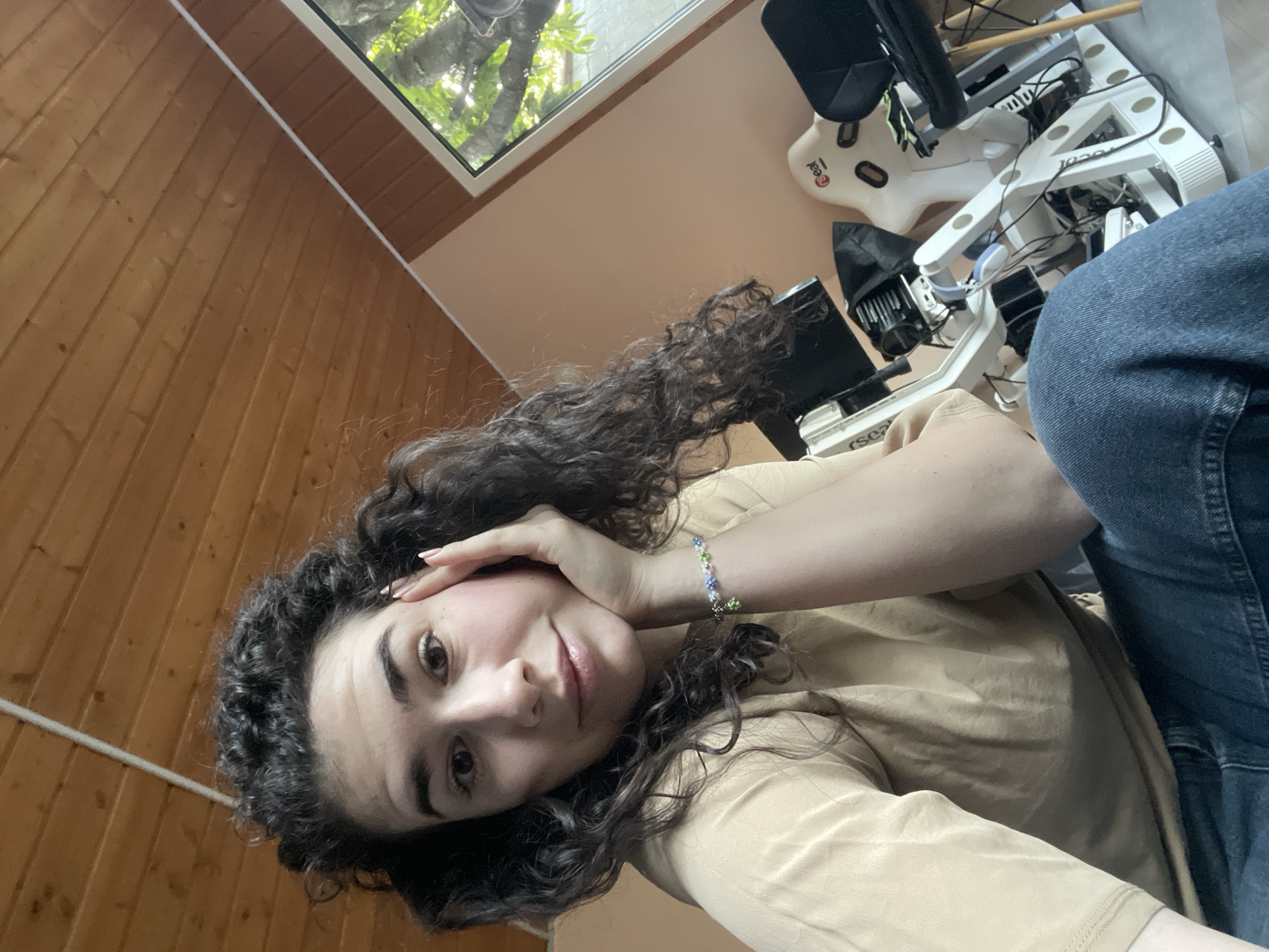 A selfie angled from the upper right shows a woman with dark, curly hair and a light complexion wearing a beige shirt and blue jeans. She has a floral beaded bracelet on her right wrist, which she is using to touch the right side of her forehead. The background is a wood-paneled wall to the left and a window at the top, through which trees are visible. To the right of the woman is a cluttered desk or table with various white and black technological objects and equipment. It is impossible to determine the location in the city of Sofia, Bulgaria, based on this image alone.