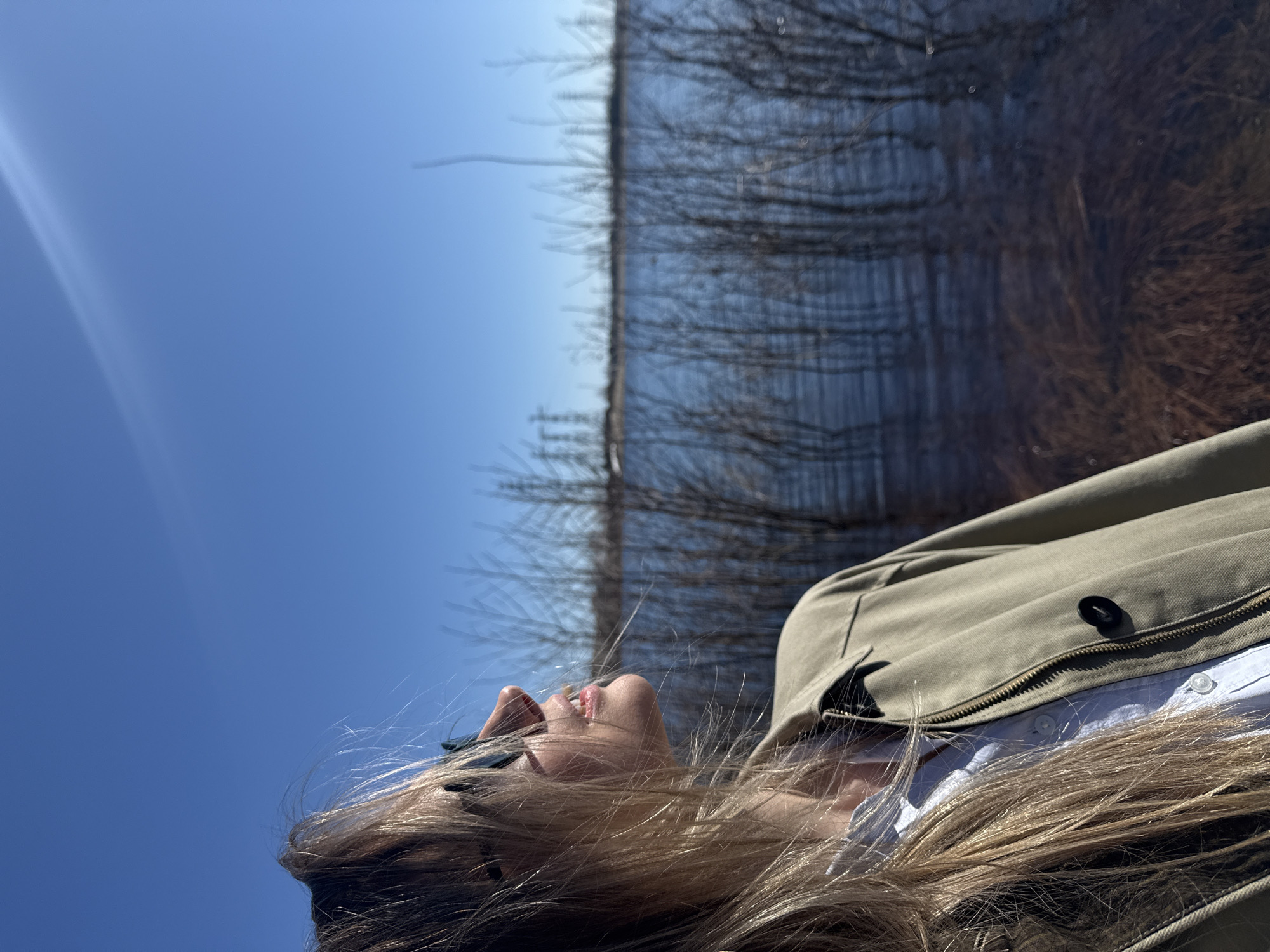 The image shows a woman lying on her back outdoors, with a clear blue sky above her and a blurred background of what appears to be water reflecting trees. She has blonde hair, is wearing sunglasses, and has an open light-colored jacket over a white button-down shirt. A zipper can be seen on her jacket. The scene is brightly lit, with a lens flare in the upper left. Based on the provided information, the location is in the city of Chaikovsky, Russia.