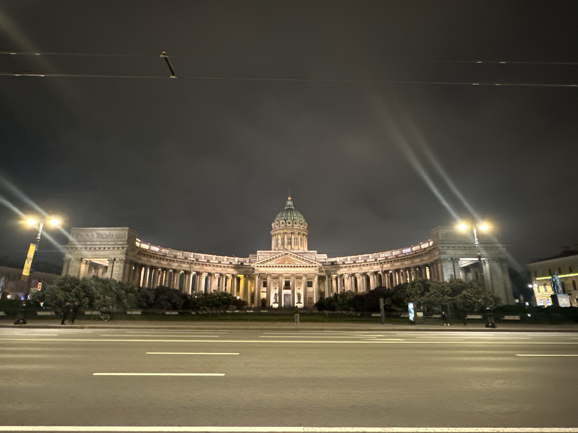 Here is a description of the image:
The image shows Kazan Cathedral in Saint Petersburg, Russia at night. The cathedral's dome and colonnades are illuminated. An empty road spans the foreground, with painted lane markings visible. Streetlights cast bright beams. A figure stands on the sidewalk. There is overcast sky in the background.