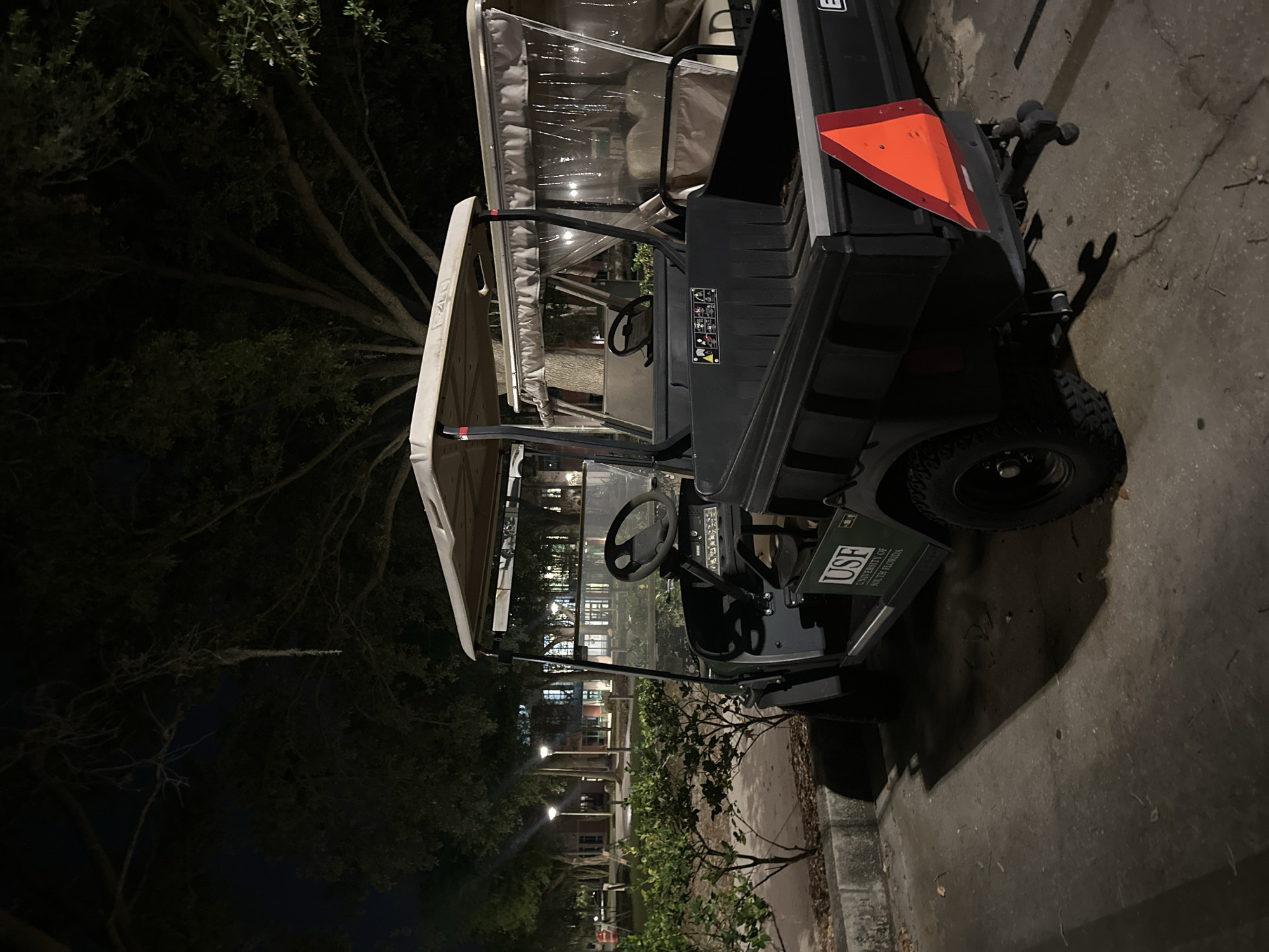 An outdoor, nighttime scene depicts a dark green utility vehicle, resembling a golf cart, parked on a concrete surface. The vehicle has a white canopy and a black body. An orange triangular slow-moving vehicle sign is affixed to its rear. On the side of the vehicle, a prominent logo reads "USF" with "UNIVERSITY OF SOUTH FLORIDA" beneath it, indicating its affiliation.
The setting is dark, illuminated by artificial light sources including streetlights and lights emanating from multi-story buildings visible in the background, seen through the vehicle's windshield. Large trees, some with visible Spanish moss, frame the left side of the image, suggesting a subtropical environment consistent with Tampa, United States of America. No individuals are present, and the vehicle appears to be stationary and unoccupied. The ground beneath the vehicle is a grey concrete path or drive. The atmosphere is quiet, typical of a campus or institutional area after dark.