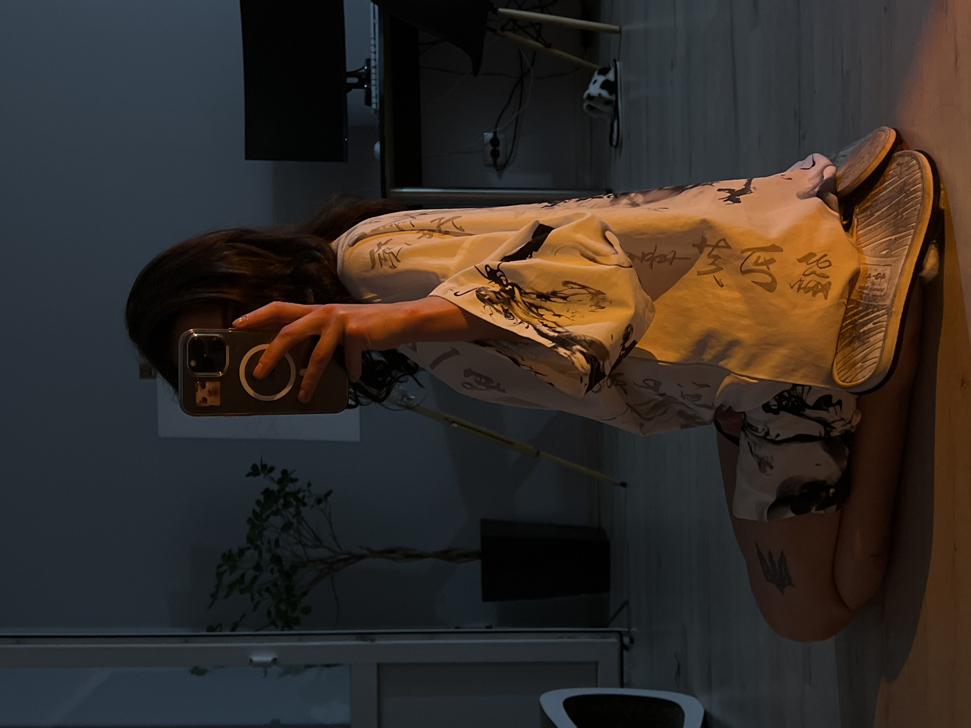A person with dark hair is sitting on the floor in a dimly lit room, taking a selfie with a smartphone. The person is wearing a light-colored robe with a dark dragon-like pattern and Chinese characters. They are wearing sandals. A potted plant is visible in the background, along with a desk with a computer monitor, and a chair. The floor is light-colored wood. The lighting in the room is warm, with a spotlight illuminating the lower half of the person. The image appears to be taken indoors, possibly in an apartment in Ternopil, Ukraine.