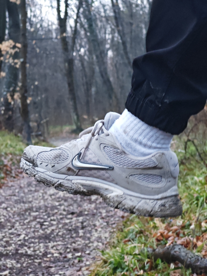 A close-up photograph captures the lower right leg and foot of an individual in a forested setting within the city of Shumen, Bulgaria. The subject wears black pants with an elasticized cuff, white ribbed ankle socks, and a light-beige or grey Nike athletic shoe. The shoe features mesh panels and a silver-toned Nike swoosh logo on its lateral side. Both the shoe, particularly the sole and front upper, and the socks display an accumulation of light-brown mud and dirt. The lower pant cuff also shows minor smudges. The foot is depicted suspended mid-air, indicative of being lifted from the ground. The immediate foreground reveals a dirt path scattered with dry brown leaves and small debris. To the right, green ground cover and additional dry leaves are present, along with a visible piece of wood or root. The background consists of a dense, leafless deciduous forest, typical of a cold season, under what appears to be an overcast sky.