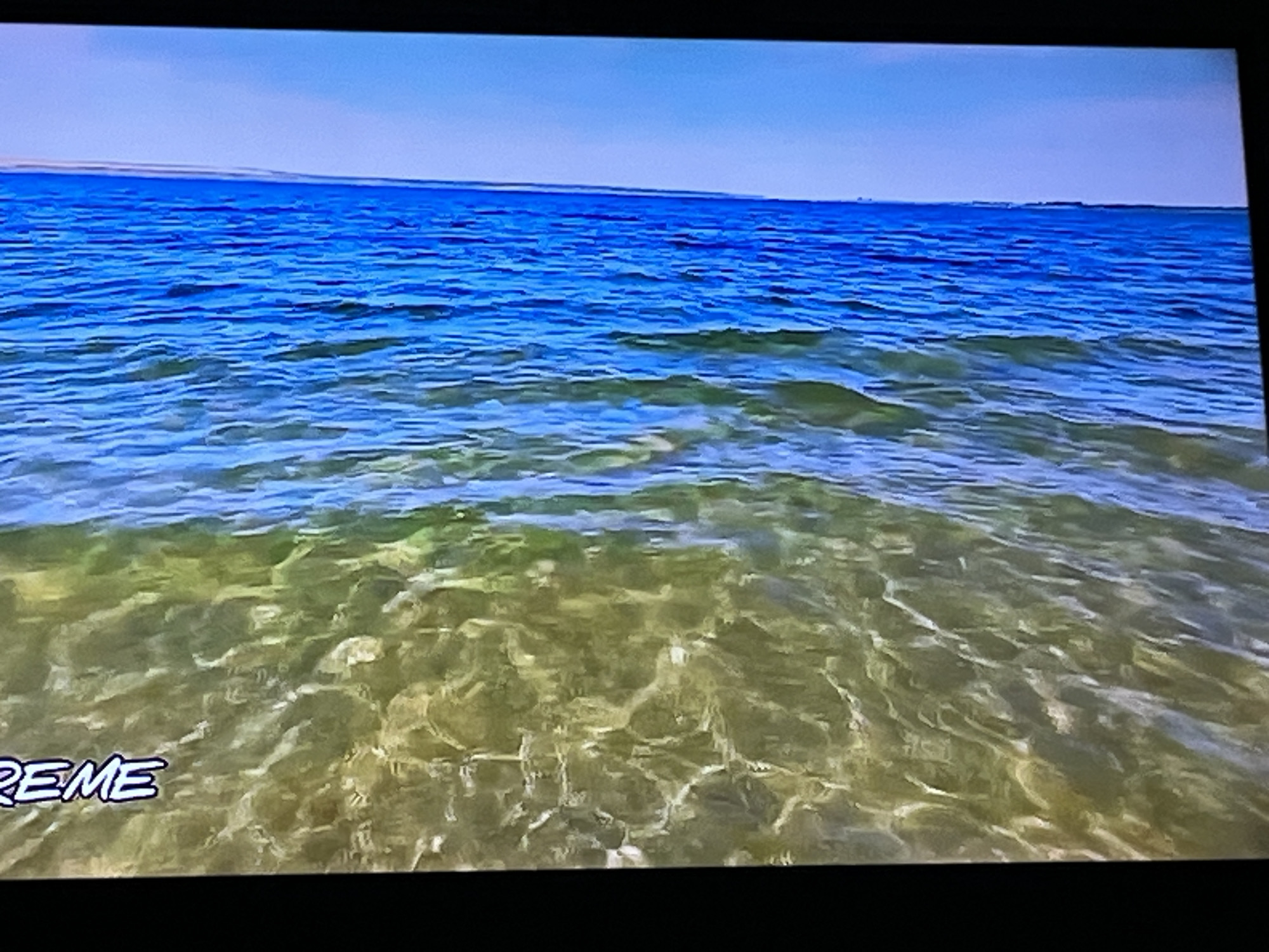 The image is a photograph of a body of water with a light blue sky above. The water is clear and shows the sandy bottom beneath. There are some white wave crests visible. In the lower left corner of the image, text that appears to say "REME" is visible. The photograph does not provide any information about the city of Kharkiv, Ukraine, or any specific location. There are no people, subjects, objects, or actions depicted in the image beyond the natural environment.