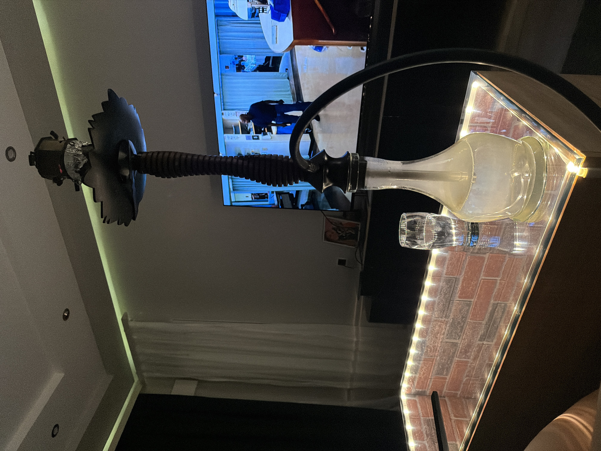 The image depicts a dimly lit indoor residential or lounge setting in Sofia, Bulgaria, characterized by modern design and ambient lighting. In the foreground, a prominent dark-stemmed hookah with a clear glass base, containing a translucent liquid and visible vapor, rests on a rectangular table. This table features a transparent top surface, underneath which a brick-patterned panel is illuminated by a perimeter of bright LED strip lights. A tall, empty clear glass stands to the right of the hookah on the table. A portion of a human limb, likely an arm or leg, is visible in the bottom left, suggesting an occupant nearby.
The midground is dominated by a large flat-screen television displaying a scene within what appears to be a hospital or clinical environment. A dark-skinned male figure, dressed in a dark blue shirt and jeans, stands in a brightly lit, blue-toned hallway, looking towards the right. Blue privacy curtains and medical equipment are visible in the background of the television screen. Below the television, a dark console holds a small framed picture on its right side.
The background reveals architectural details including a light-colored wall with white sheer curtains covering a window or doorway on the left. The ceiling features recessed circular lights, one appearing unlit and another dimly illuminated. Green-toned LED strip lighting runs along a ceiling cove, providing indirect illumination to the upper wall. The overall ambiance is subdued and intimate, contrasting with the bright, cool tones emitted by the television display.