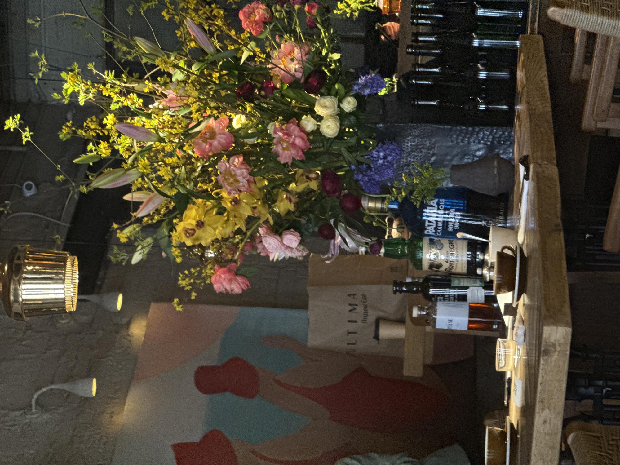 The image, rotated 90 degrees counter-clockwise, depicts an inviting indoor scene, likely a bar or restaurant in Yekaterinburg, Russia. The central focus is a large, vibrant floral arrangement overflowing with various types of blossoms. Yellow forsythia-like branches, daffodils, and lilies are prominent, alongside pink peonies and lilies (some still in bud), white roses, purple hyacinth-like clusters, and deep burgundy flowers.
The floral display rests on a warm-toned wooden counter or bar. To its right, several bottles of spirits are lined up, including one clearly labeled "MONTENEGRO" and another blue bottle with "PATATATA CHAMP OIS SOLES." A brown ceramic cup with a saucer and various clear glasses, some containing liquid, are also present on the counter.
In the background, a large, abstract mural on the wall features stylized human-like figures in shades of red, blue, and beige. Illumination comes from golden-hued pendant lamps and spotlights accentuating the mural. A security camera is discreetly mounted on a wall section in the upper left. A paper bag with "ULTIMA" and "Яндекс Еда" (Yandex Food in Cyrillic) is visible near the flowers, confirming the Russian context.
While no people are distinctly visible, partial arms or hands suggest human presence, likely patrons or staff. The artificial lighting indicates an evening or nighttime setting. The combination of the elaborate flowers, sophisticated drink selection, and artistic mural creates a lively and upscale ambiance.