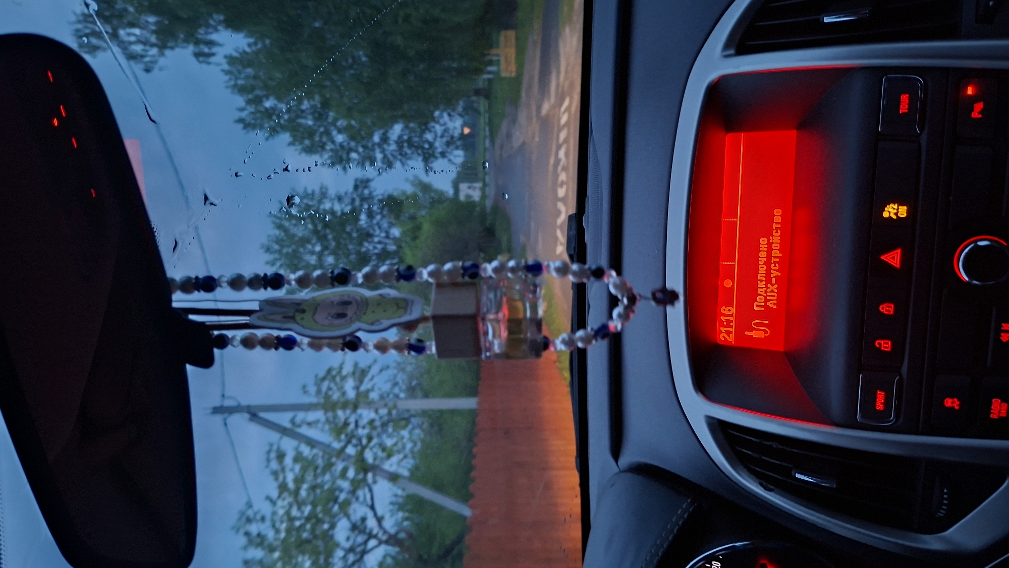 The image depicts the interior of a car, likely an Opel Astra, taken from the driver's perspective. The car is located in or near Забалоцкі сельскі Савет, Belarus, as evidenced by the reflection in the windshield showing a rural road with sparse housing and roadside vegetation typical of the area. The time displayed on the dashboard indicates 21:16 (9:16 PM). The radio display is lit in red, showing the text "Подключено AUX-устройство" which translates to "AUX device connected." There is a string of beads with a small cartoon pendant hanging from the rearview mirror. The external view through the windshield suggests twilight or early evening with visible raindrops, indicating recent or ongoing rain. The road ahead is wet and illuminated by the car's headlights. The red roof of a building or fence is seen in the lower part of the windshield. The car's dashboard buttons are illuminated in red, including hazard lights and other controls.