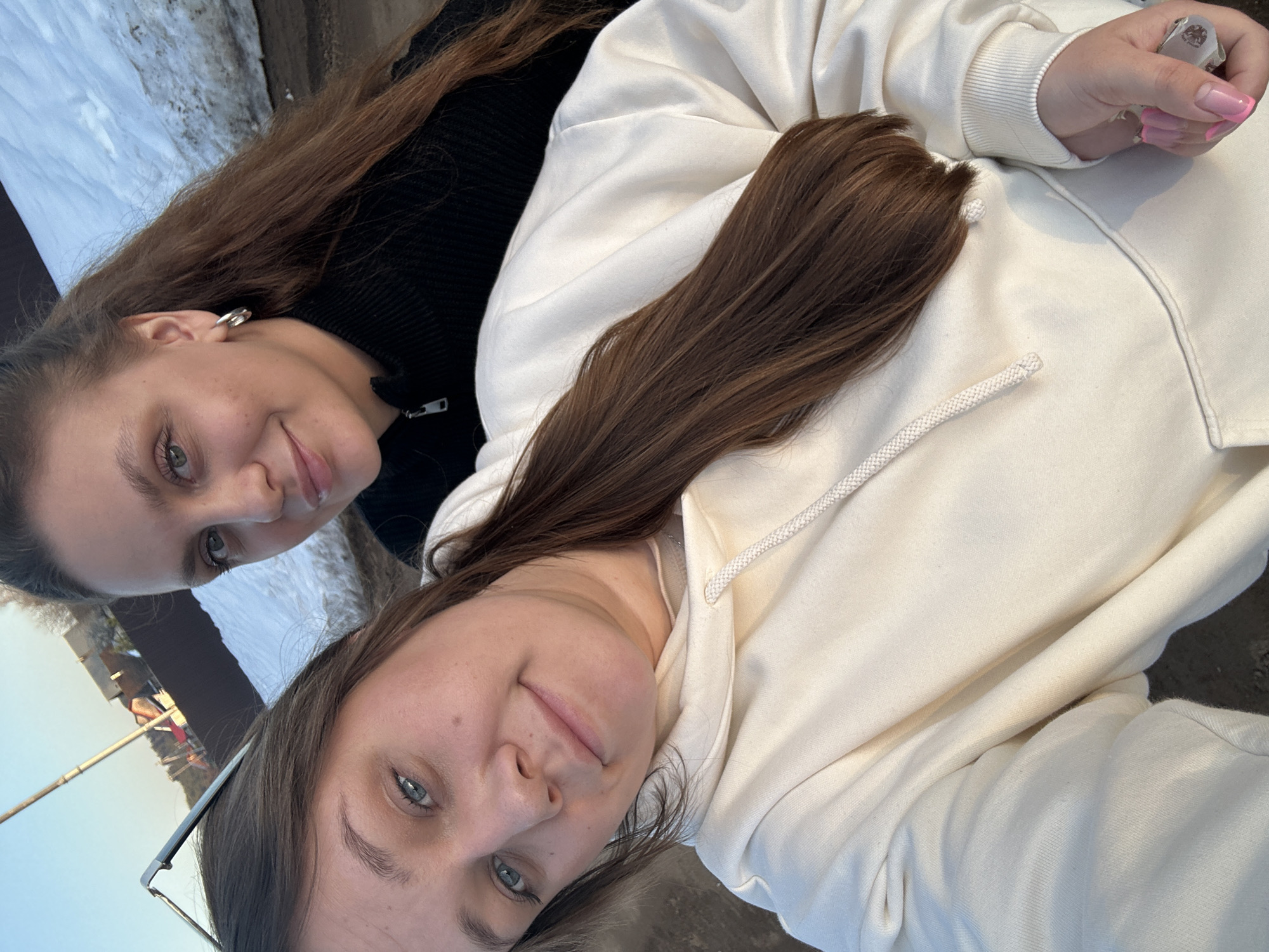 Two fair-skinned women are captured in a selfie, presumably in the city of Ольховка, Russia, given the context. One woman, positioned at the top of the frame, wears a black top and has long, light brown hair. The second woman, at the bottom, wears glasses perched on her head and a white hoodie. Both women look towards the camera with neutral expressions. The background suggests an outdoor setting, featuring snow, a body of water, and distant structures. The angle suggests a personal snapshot. A manicured hand, wearing a silver ring with pink nails, is visible on the right side of the image.