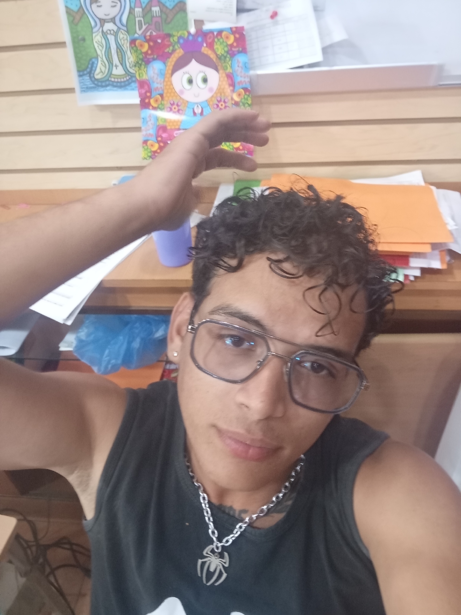 A selfie taken by a young adult male with curly dark hair and eyeglasses. He is wearing a dark tank top and a silver chain necklace with a spider pendant. His right arm is raised above his head. The background shows a wooden-paneled wall with two cartoon drawings affixed to it, and a cluttered desk with papers, a purple cup, and a blue plastic bag. The location is likely indoors, potentially in a home or office space. There is no definitive evidence within the image to confirm it is specifically in Juan Griego, Venezuela.