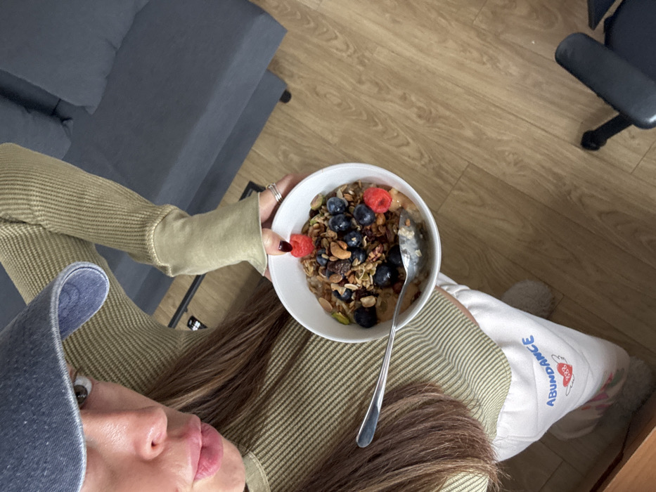 A female subject is depicted from an overhead perspective in an indoor setting within the city of Unknown, Unknown. She wears a blue denim baseball cap, an olive-green ribbed long-sleeve top with extended cuffs, and white sweatpants featuring "Abundance 333" text with a heart and wings graphic. Her right hand, adorned with rings and dark nail polish, holds a white bowl containing a meal composed of a base topped with blueberries, raspberries, pistachios, cashews, and granola; a silver spoon rests within the bowl. The subject's face is partially visible, with one eye directed at the camera and lips slightly pursed. The background features light brown wooden flooring, a partial view of a dark grey fabric sofa, and a black wheeled chair.