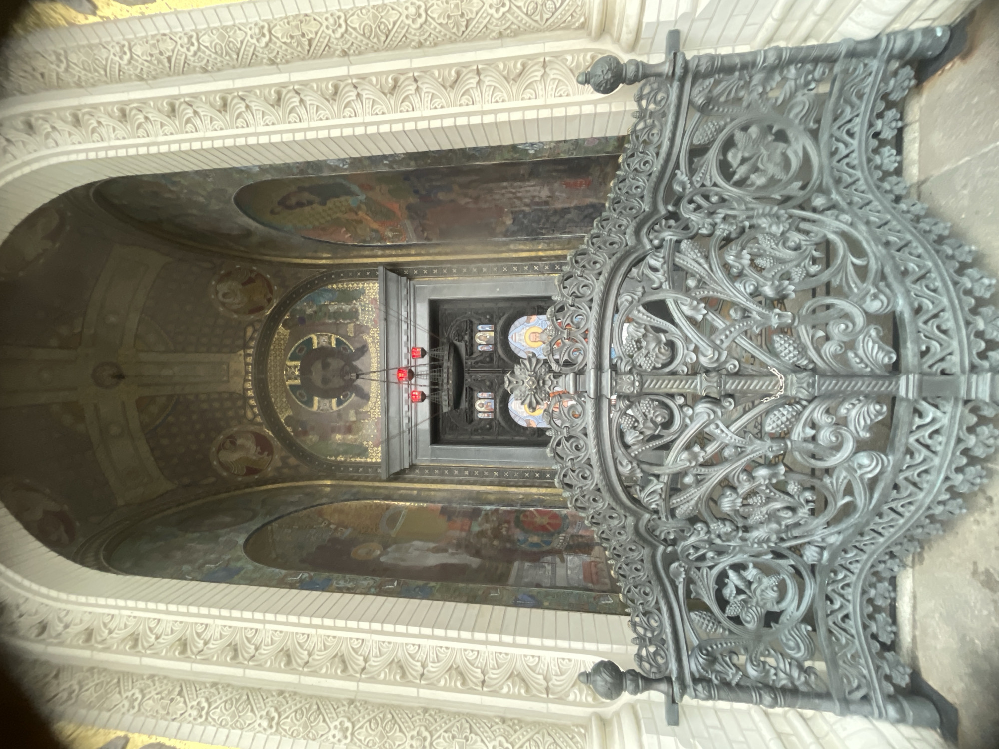 The image shows the ornate entrance to St. Nicholas Naval Cathedral in Kronstadt, Russia. A dark, intricately designed metal fence with decorative scrollwork, grape clusters, and floral motifs occupies the foreground, partially obscuring the view of the cathedral's entrance. Behind the fence, a large, arched entryway is visible, adorned with colorful mosaics and murals. A central arched doorway with a dark door is flanked by vertical panels featuring religious figures. Above the door, a circular mosaic depicts a face, possibly Jesus Christ, surrounded by gold leaf and intricate patterns. Red lights hang from a fixture above the entrance. The exterior walls of the entryway are decorated with white relief carvings in a repeating geometric and floral pattern. The overall impression is one of grandeur and religious significance.