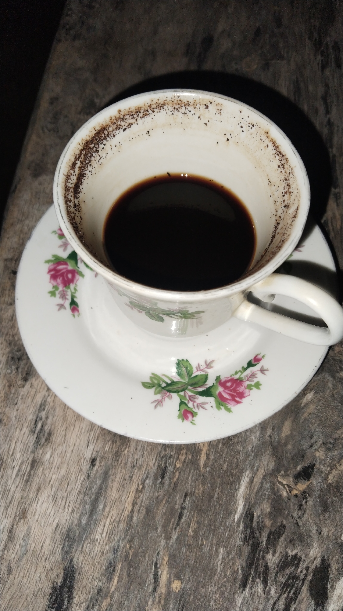 A close-up, overhead shot shows a white ceramic coffee cup and saucer decorated with pink roses and green leaves. The cup contains dark coffee, and the rim is stained with coffee grounds. The cup and saucer rest on a rustic, weathered wooden surface. The lighting is dim, creating shadows around the objects. There are no people present in the image. The scene is likely indoors or outdoors, but the specific location in Buduan, Indonesia cannot be determined from the visual information provided.