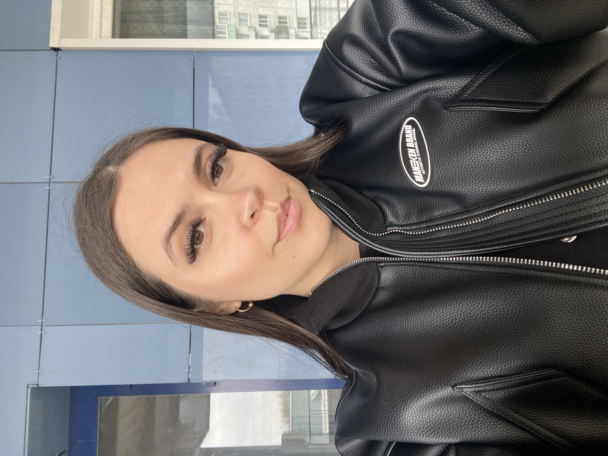 A young woman is featured in a close-up, rotated selfie, presenting a head-and-shoulder view. She has long, dark brown hair parted in the middle and styled straight, framing her face. Her brown eyes are accentuated by noticeably long, dark eyelashes, and she wears light makeup including a natural-toned lip color and subtle blush. A small gold hoop earring is visible in her left ear. She maintains a neutral to slightly smiling expression, looking directly towards the camera.
She is dressed in a black faux leather jacket with a textured, pebbled finish, worn over what appears to be a black hooded top or sweater. The jacket features a prominent silver zipper down the front and ribbed cuffs. On her right shoulder, an oval white patch with black text is visible, reading "MANEKEN BRAND" at the top and "AUTHENTIC OVERSIZE APPAREL" below it.
The background consists of a modern building exterior, characterized by large, light blue rectangular panels or tiles, some with visible seams. Reflections in the windows behind her show architectural details of other buildings and hints of a possibly snowy or cold environment. The lighting suggests it is daytime. The setting appears to be urban, consistent with the context of Chaikovsky, Russia.