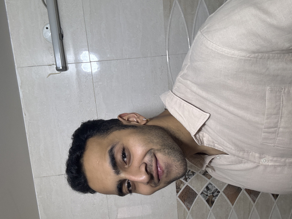 A man with dark hair, stubble, and an earring in his left ear smiles at the camera. He is wearing a light tan collared shirt. The background is a tiled wall with varying shades of white and brown patterns. A light fixture is mounted on the wall behind him. The photo has been rotated, and the man is positioned on his side.